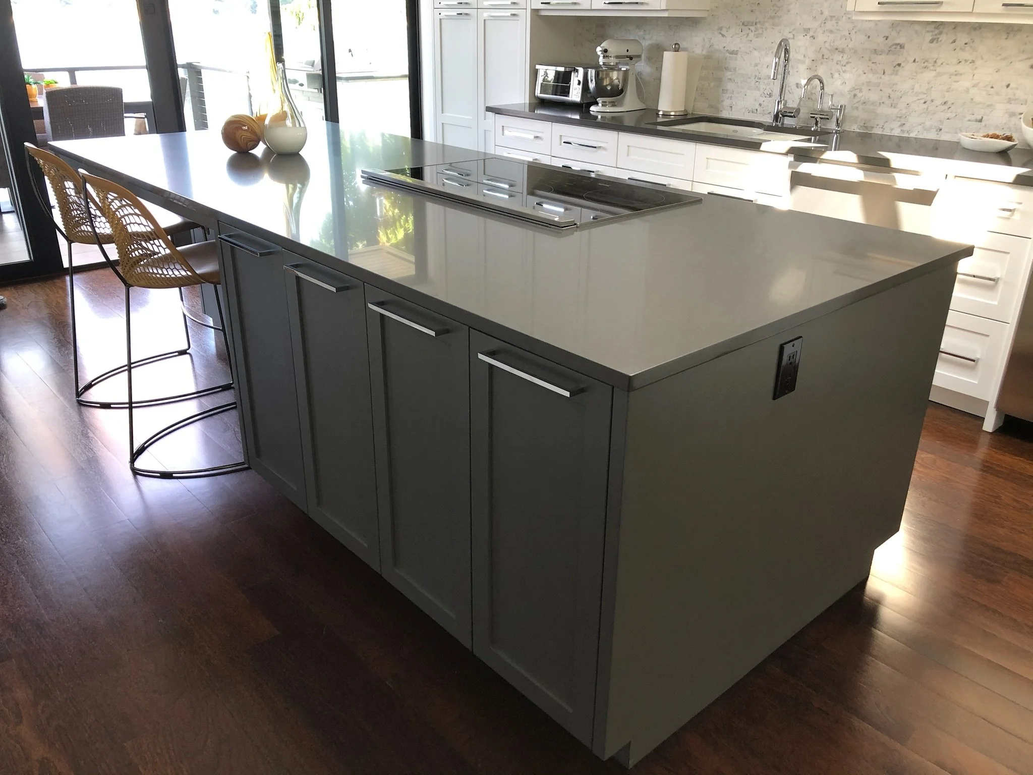 Modern kitchen island with gray cabinets, a white countertop, and barstools, located in a bright kitchen with white cabinets, stainless steel appliances, and hardwood floors.