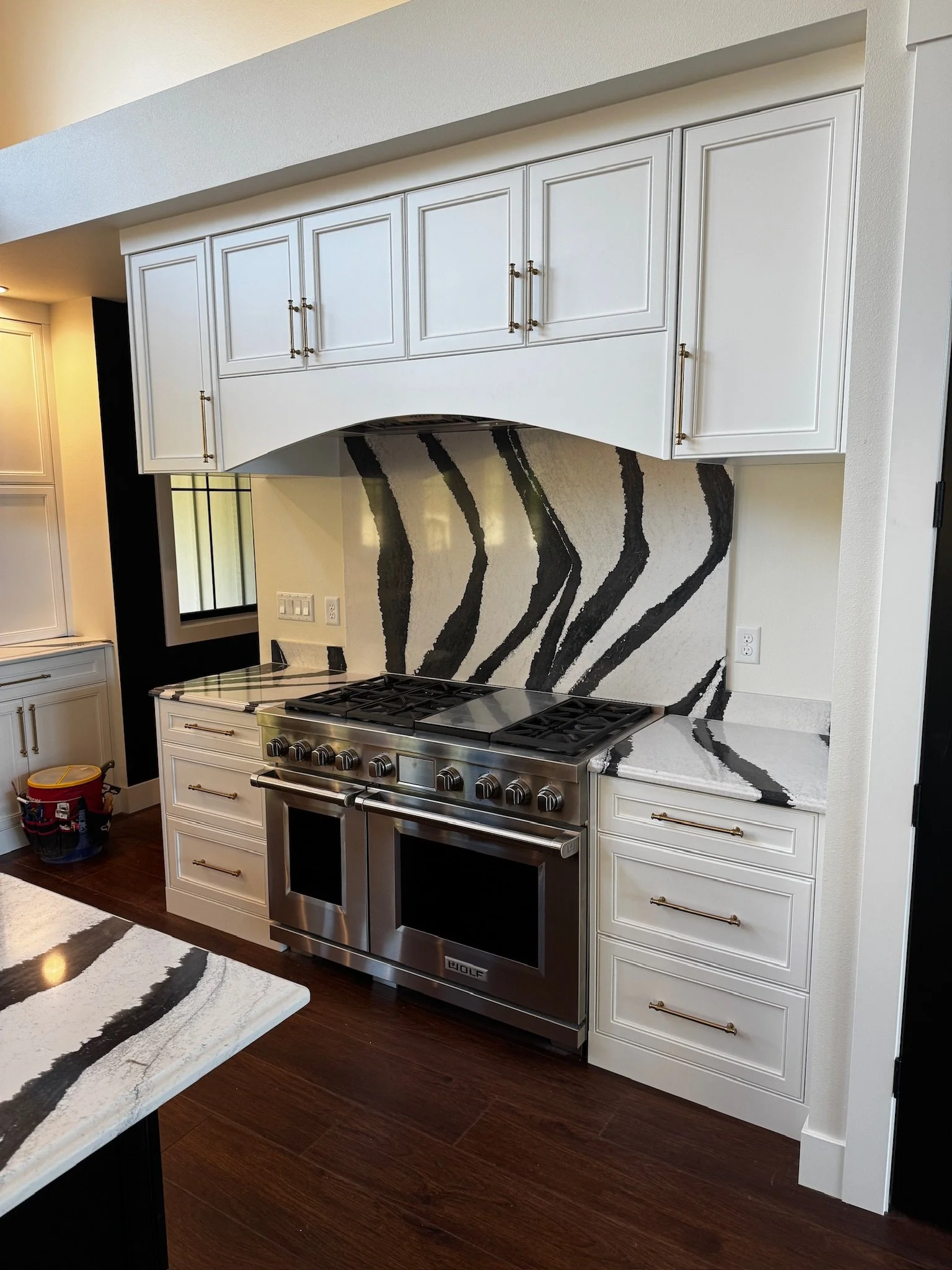 White kitchen cabinets with gold handles above a stainless steel Wolf stove with a marble countertop and a zebra-striped marble backsplash.