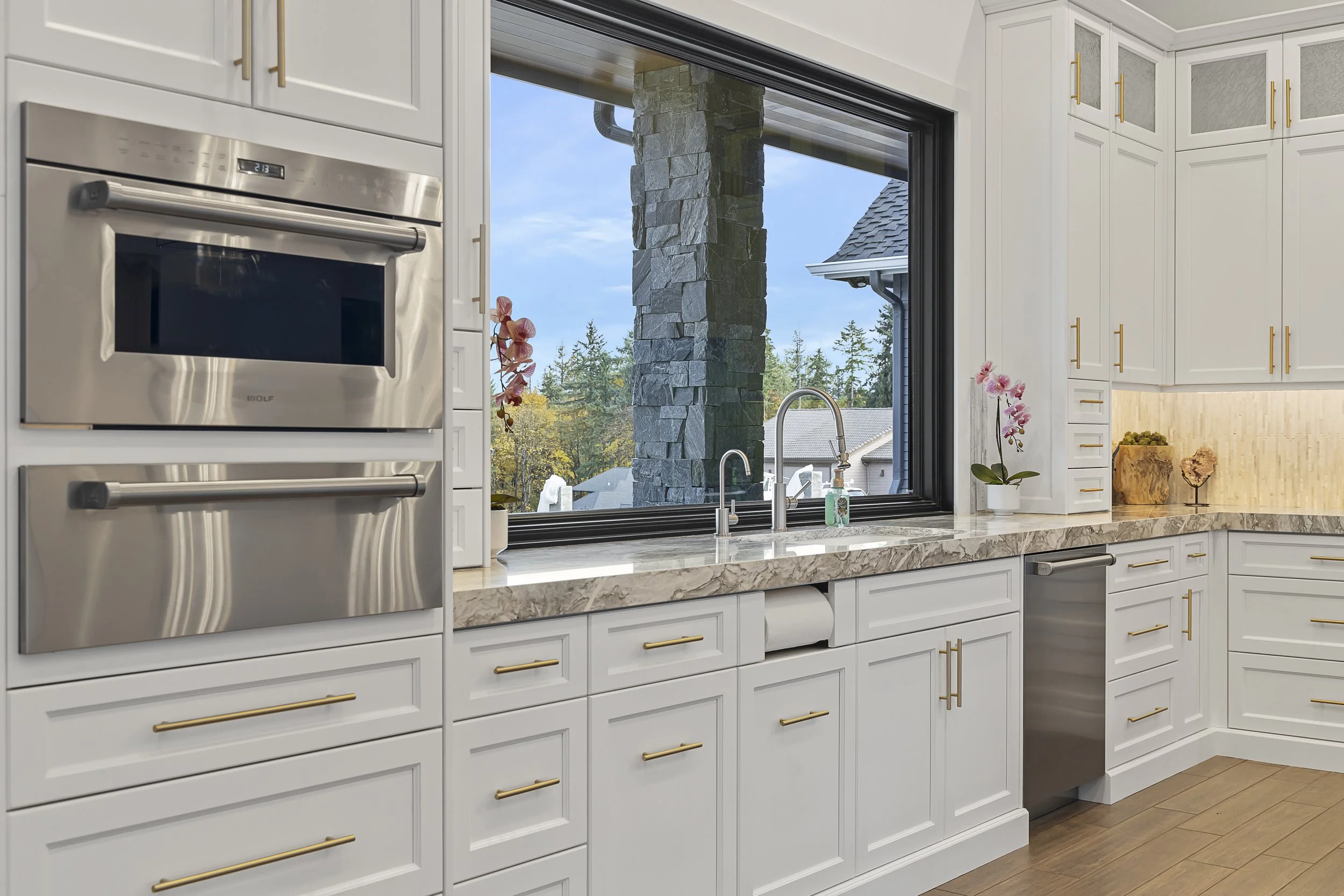 Modern kitchen with white cabinets, marble countertop, stainless steel appliances, large window showing backyard and trees, pink orchids, and wood flooring.