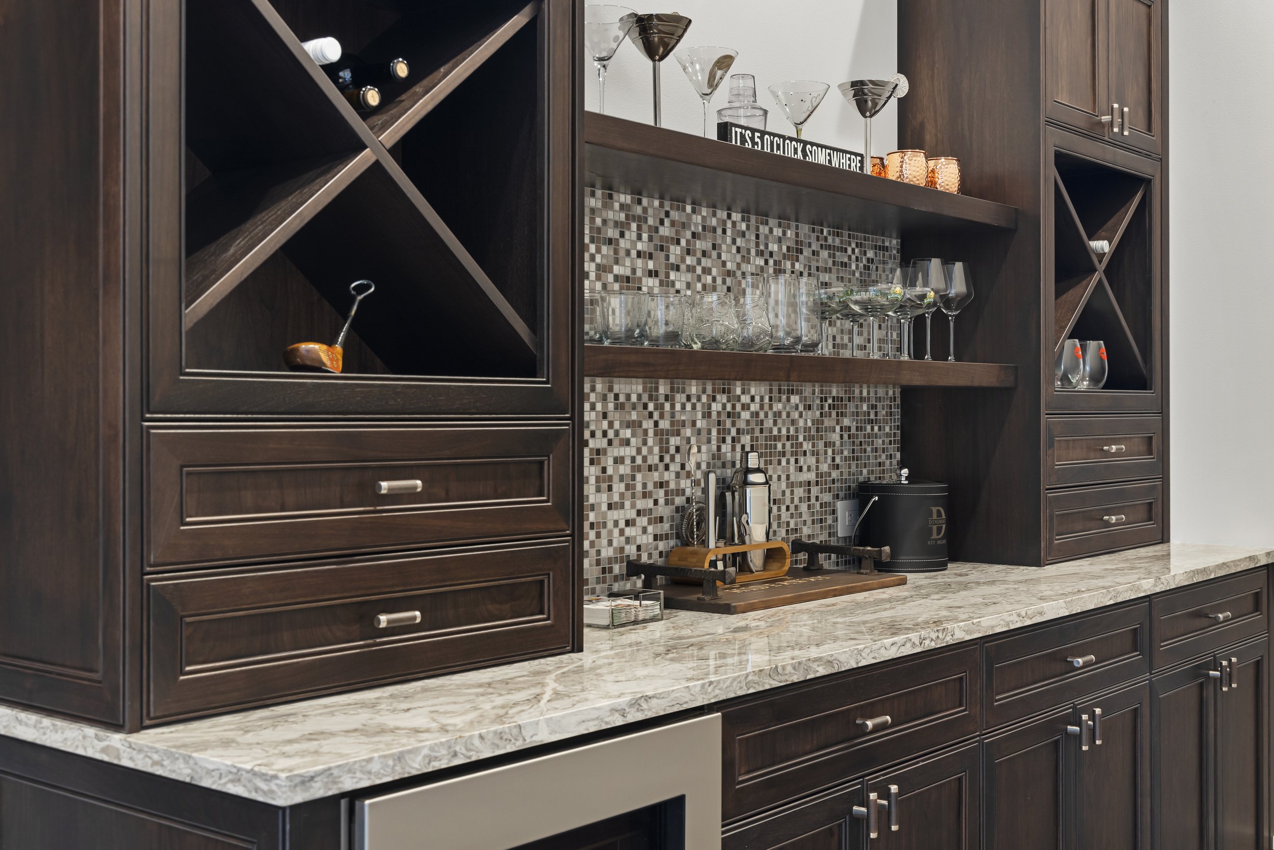 Home bar with dark wood cabinetry, a marble countertop, and glassware on open shelves, including martini, wine, and shot glasses, with a mosaic tile backsplash.