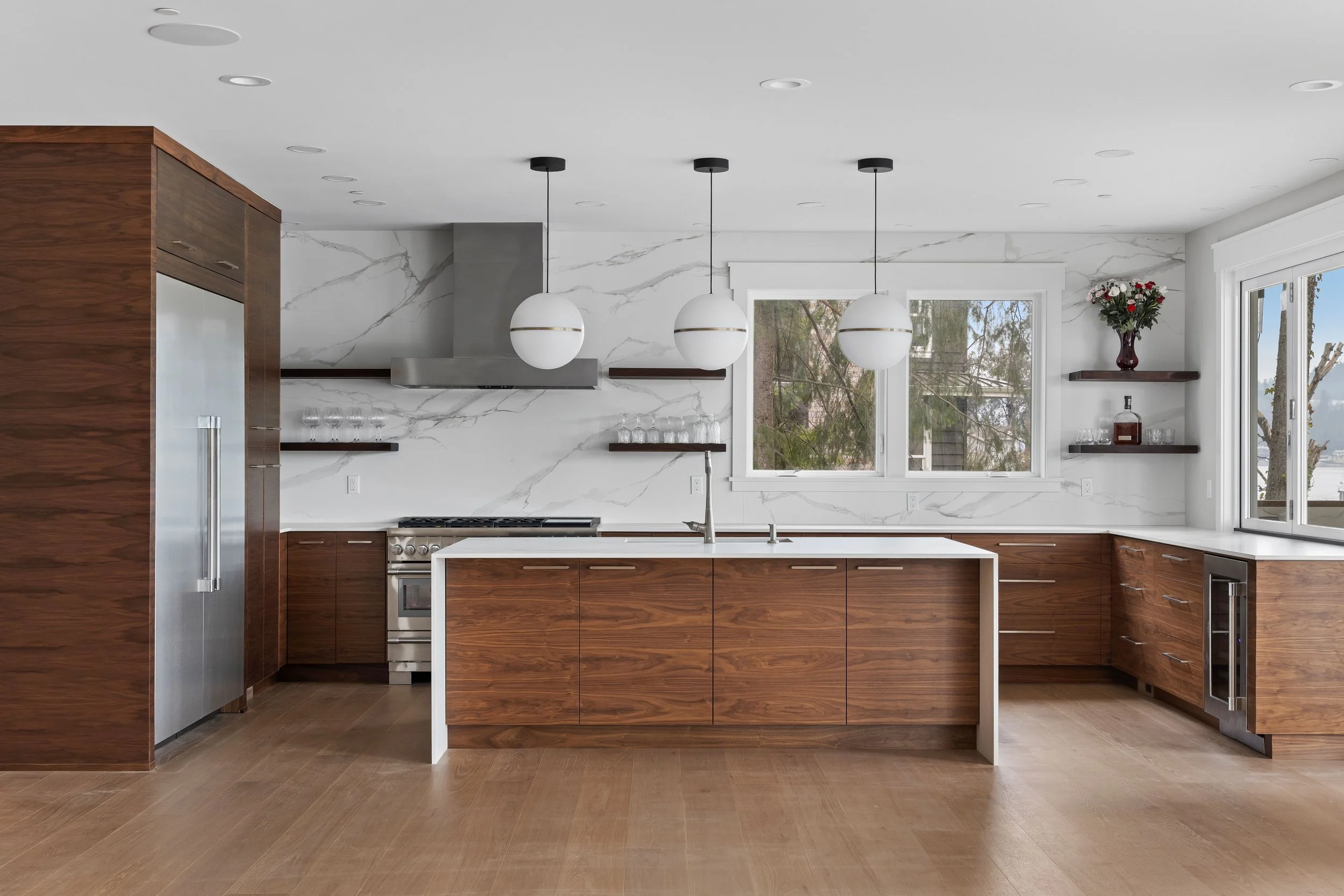 Modern kitchen with white marble backsplash, wooden cabinets, island with white countertop, stainless steel appliances, and large windows with a view of trees.