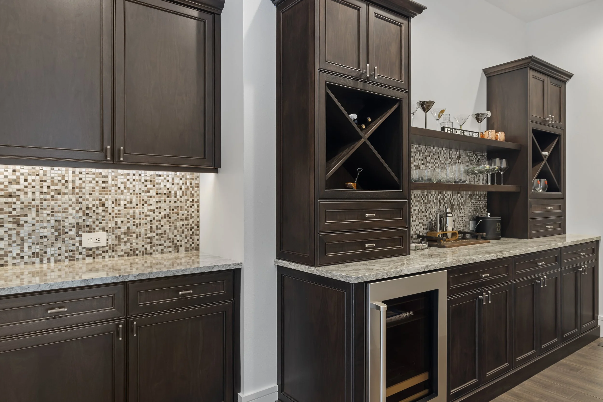 Dark wood kitchen cabinets with granite countertops, mosaic backsplash, and built-in wine storage and glassware display.