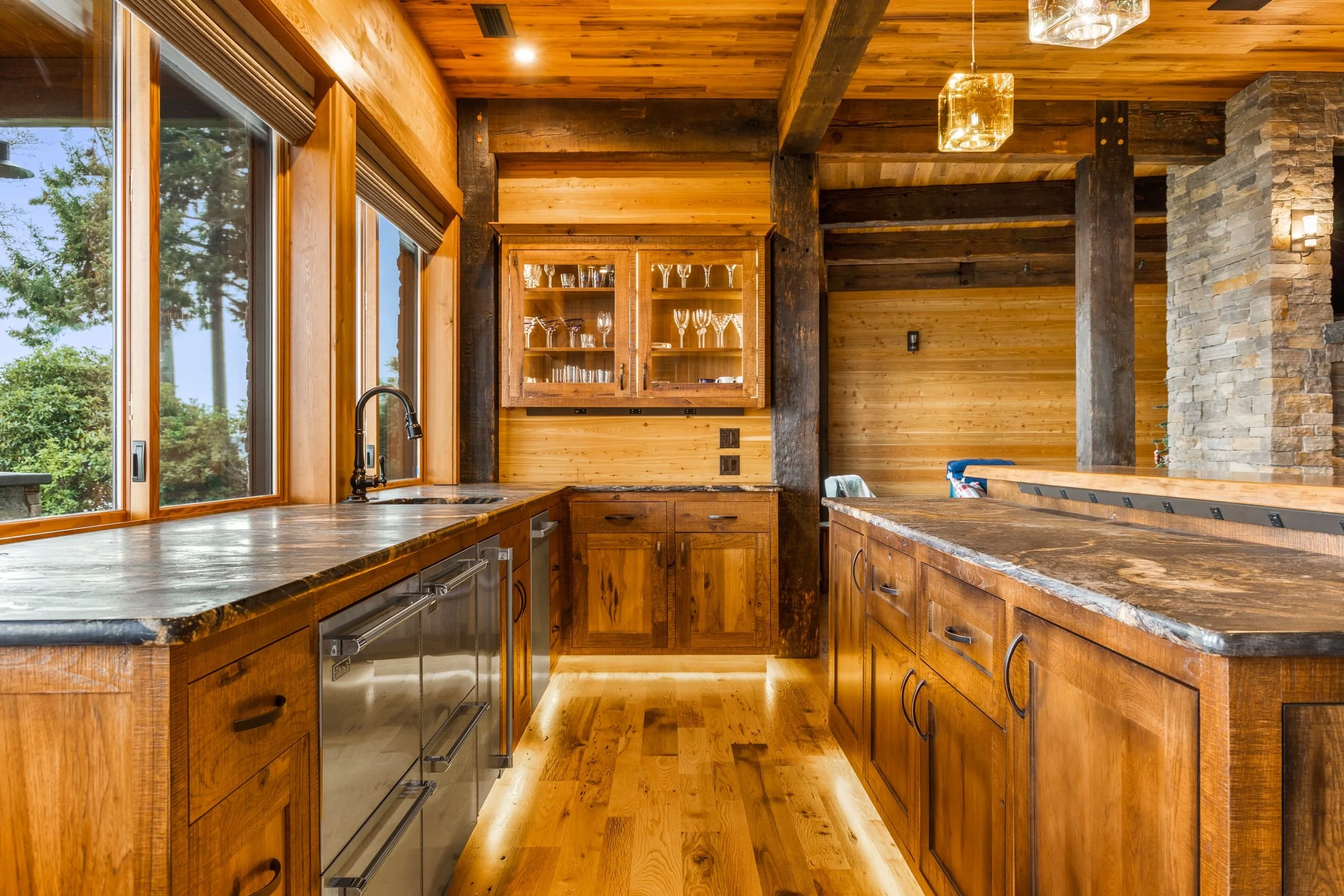 Rustic kitchen with wooden cabinets and countertops, large windows with a view of trees outside, stone fireplace, and warm lighting.