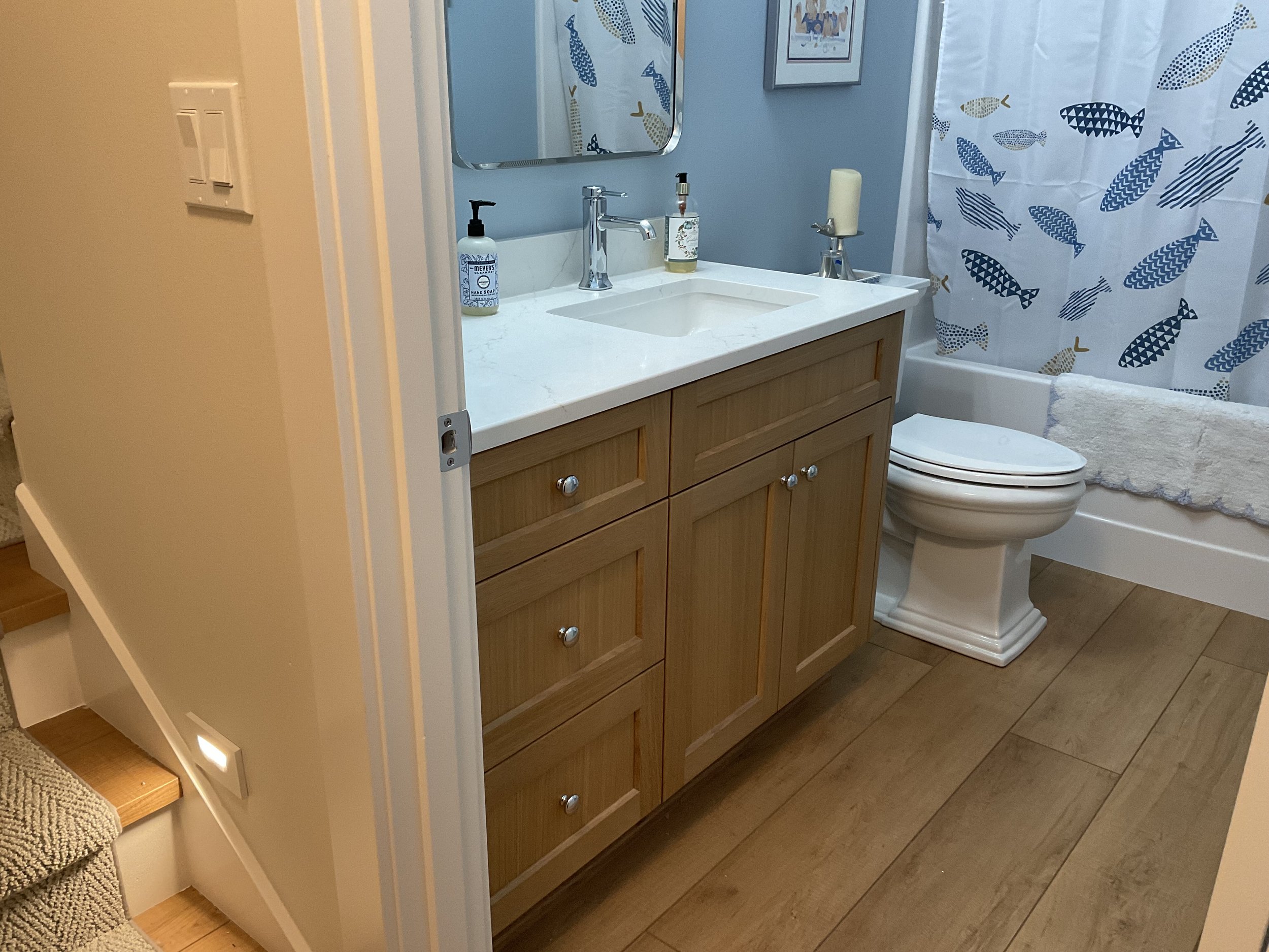 A bathroom with a wooden vanity, white countertop, soap dispenser, mirror, toilet, and a fish-patterned shower curtain.