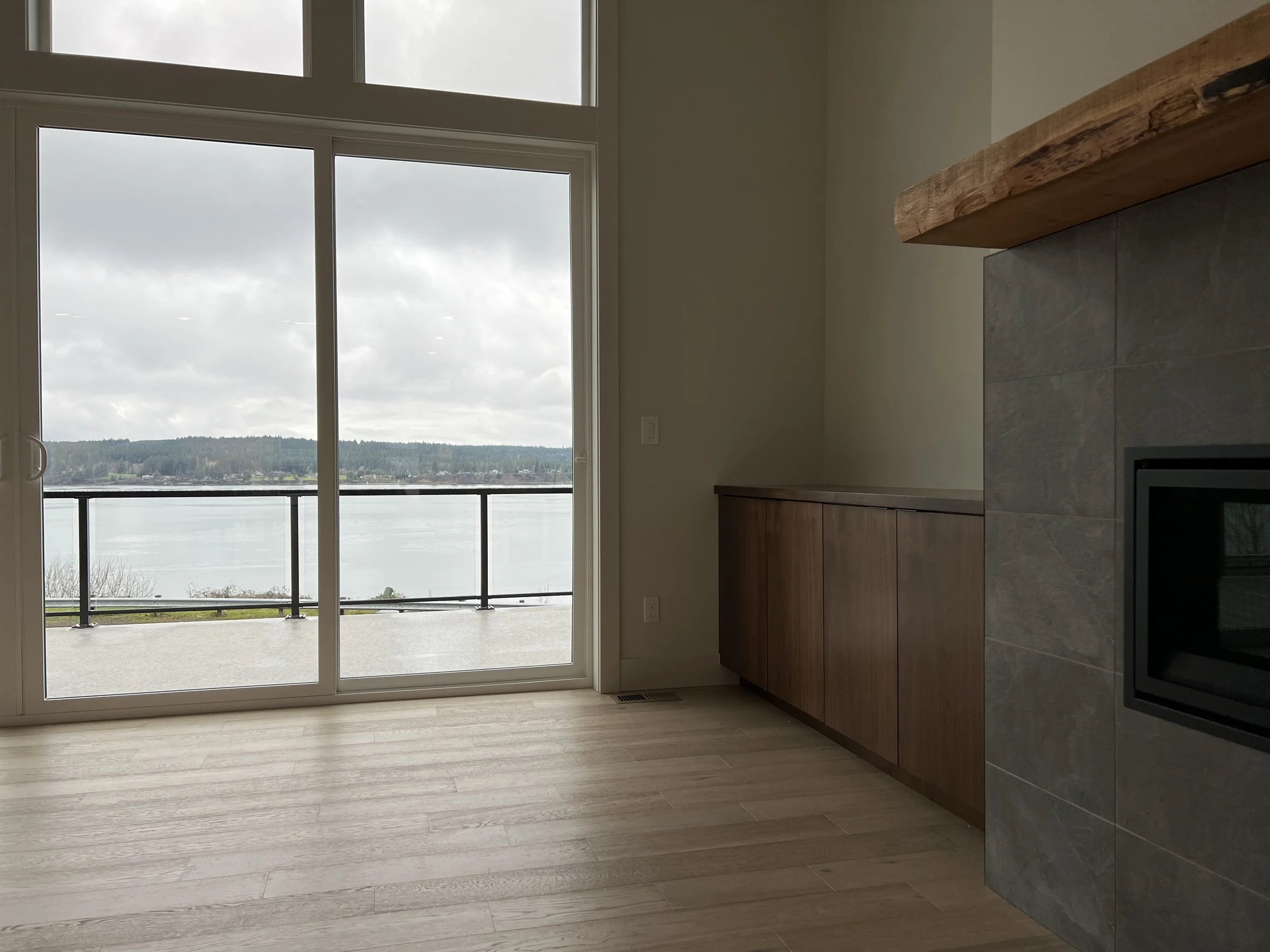 Living room with sliding glass door opening to a balcony overlooking a water body and forested landscape, with a fireplace on the right and wooden flooring.