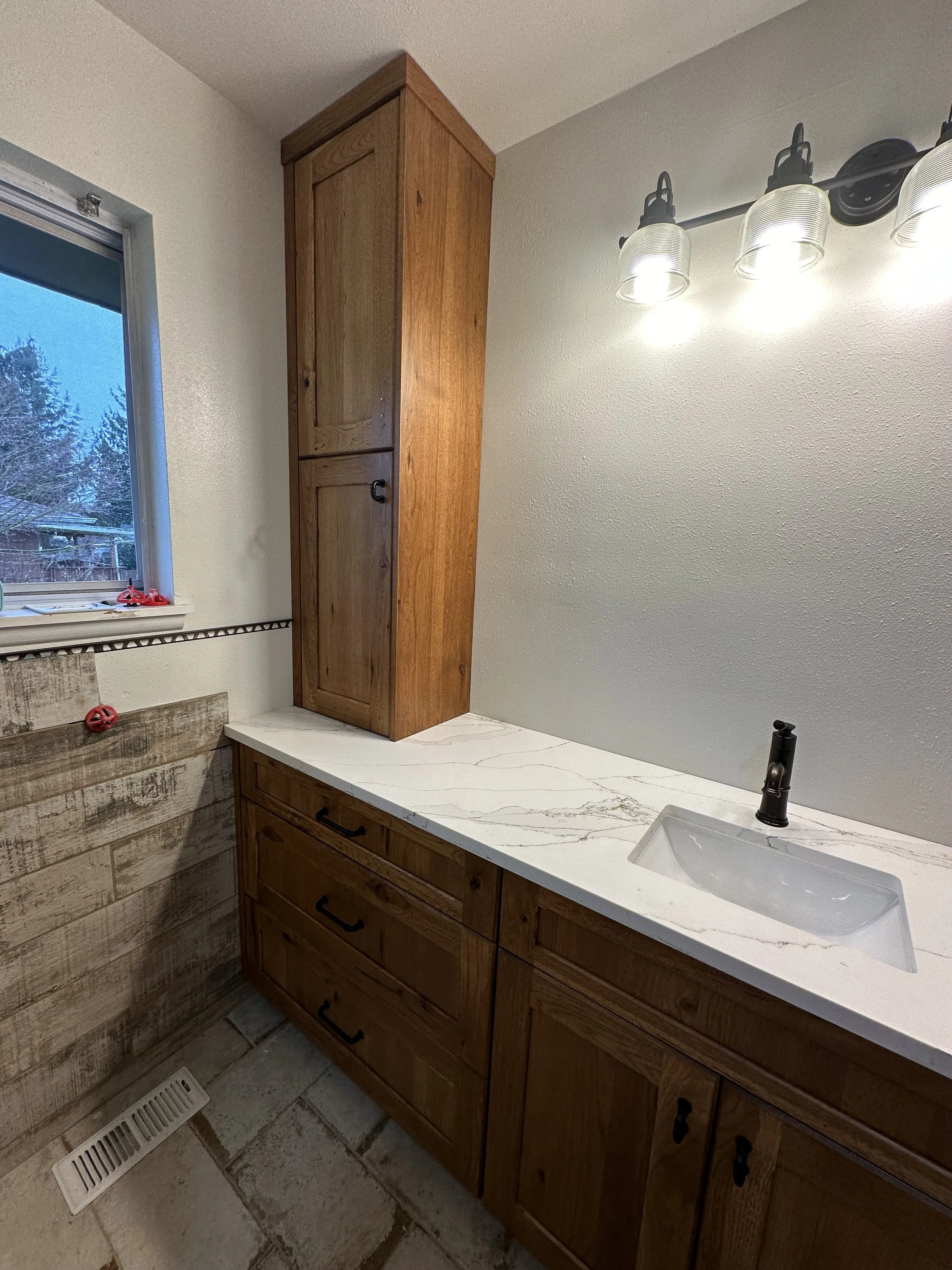 Bathroom vanity with wooden cabinets, a marble countertop, a small white sink, a black faucet, and a set of four exposed light fixtures on the wall. There is a window on the left with a view of a fence and trees. Part of a wooden wall and a floor ven