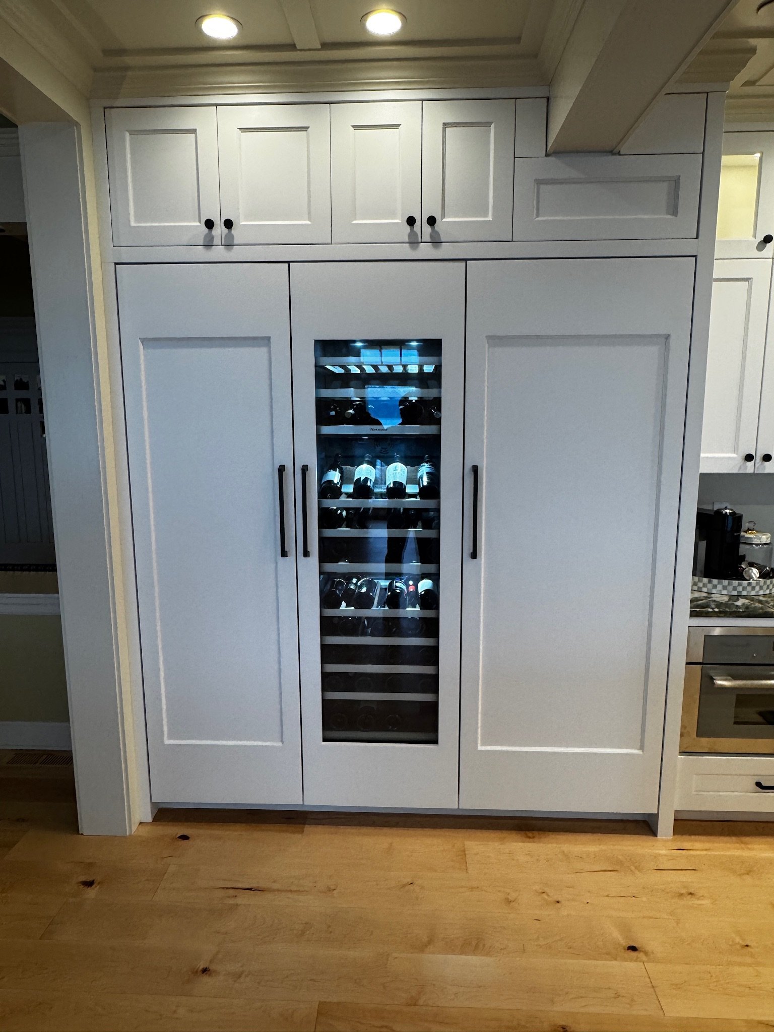 Built-in white cabinets with a glass-front wine cooler in the center, in a kitchen with a wooden floor.