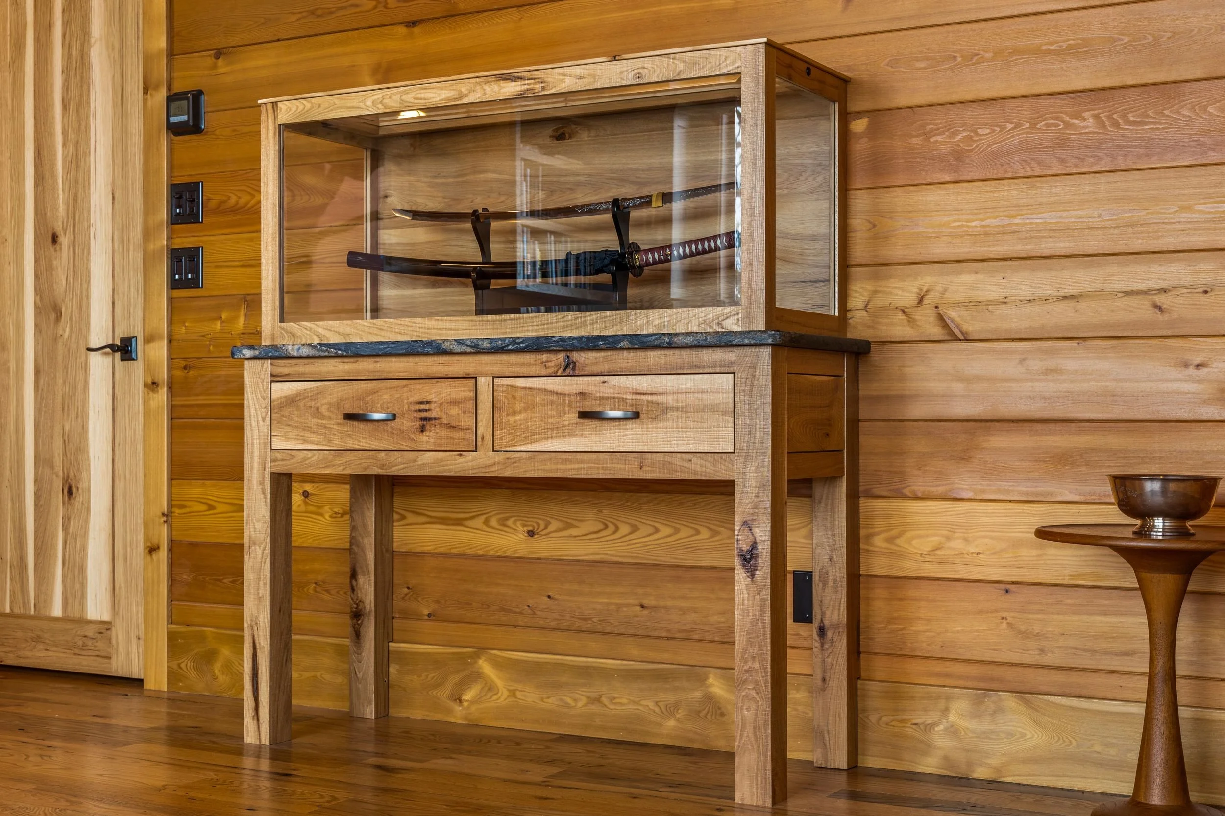 Display case with a sword inside, on a wooden stand, located against a wood-paneled wall.