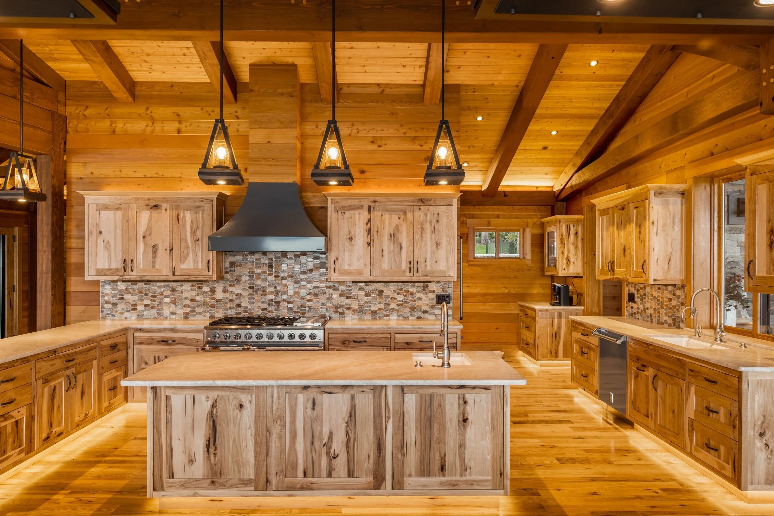 A spacious kitchen with wooden cabinets, a central island with a sink, a stovetop with a stainless steel range hood, pendant lighting, and multiple windows letting in natural light, all featuring wood paneling on the walls and ceiling.