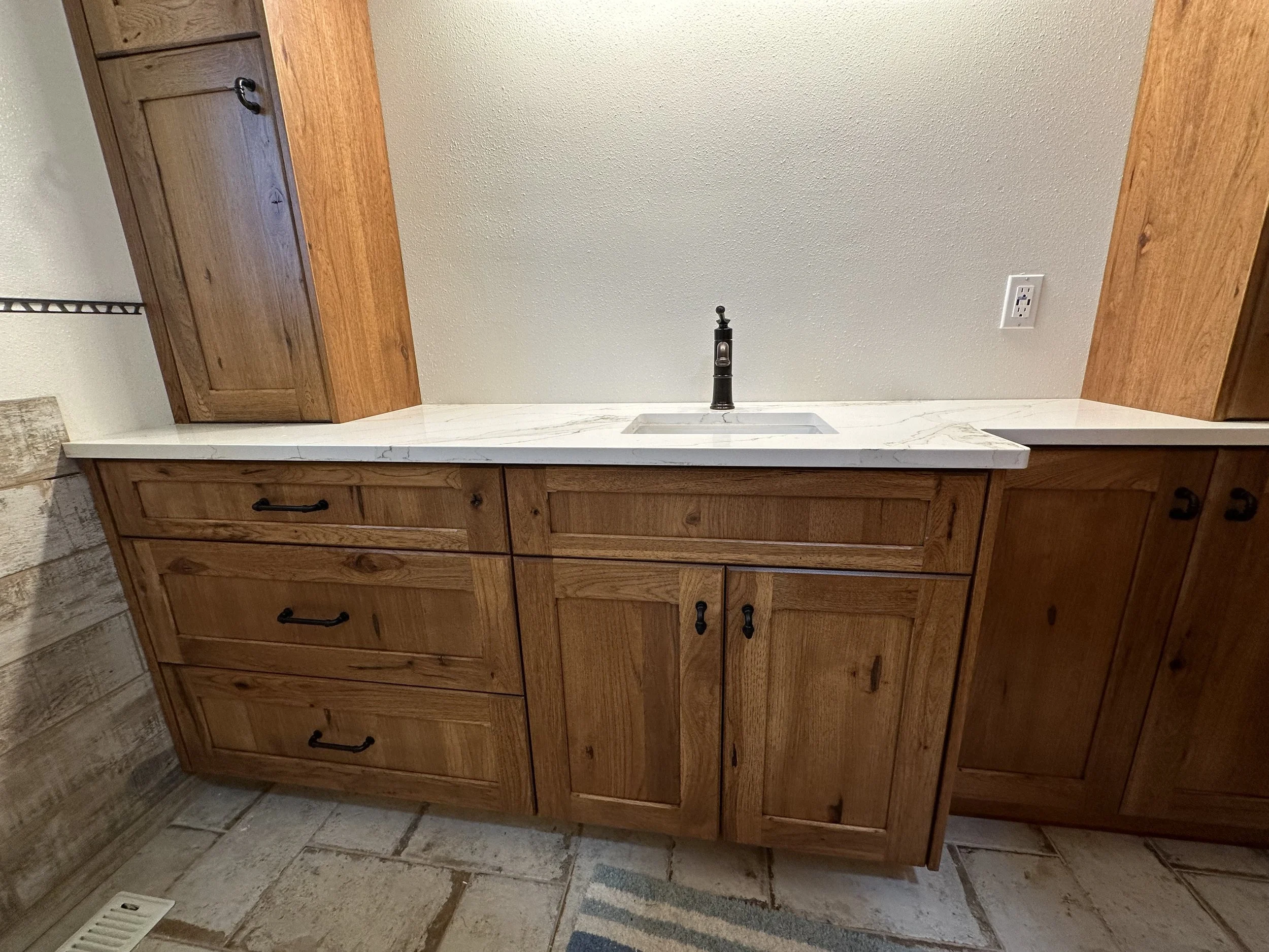 Wooden kitchen cabinet with a marble countertop, a small sink, and a black faucet, against a light-colored wall with a power outlet and nearby cabinetry.