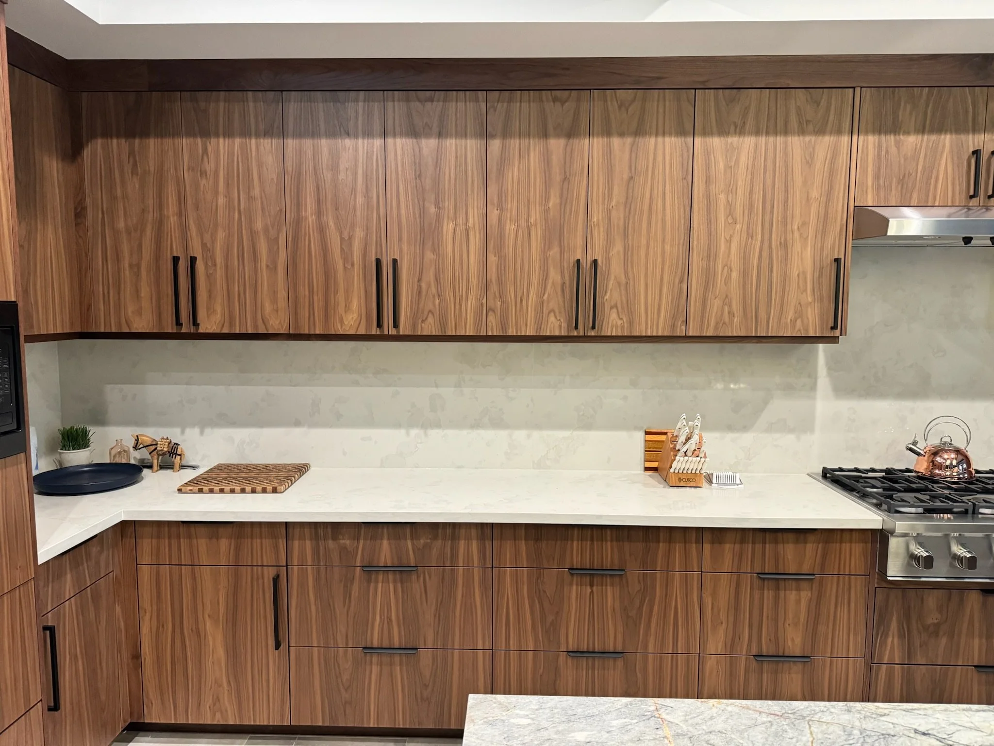 Modern kitchen with wooden cabinets, white countertops, a stove with a copper kettle, a cutting board, and small decorative items.