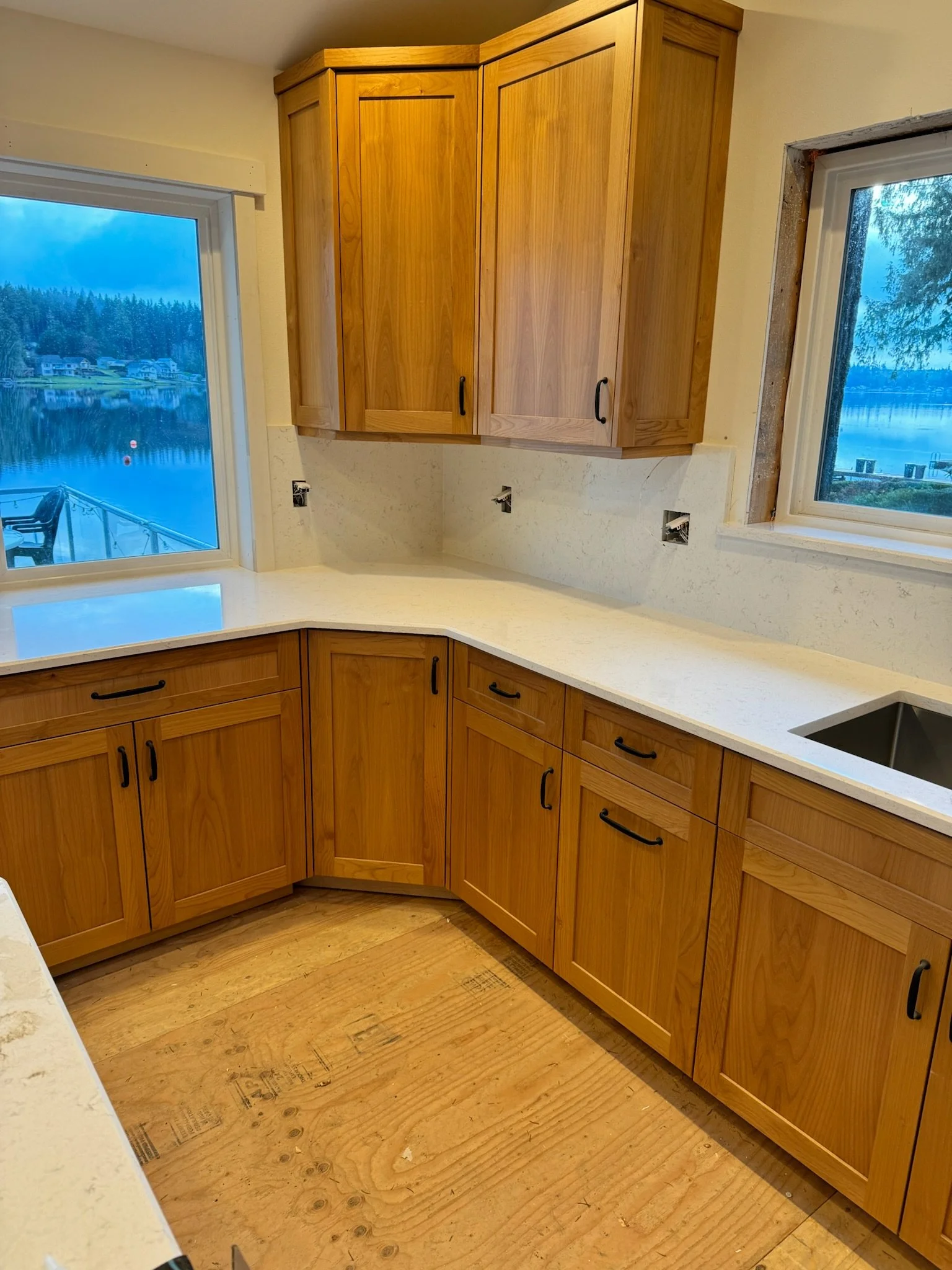 Kitchen with wooden cabinets, white countertops, and two windows showing a lakeside view, with an unfinished floor.