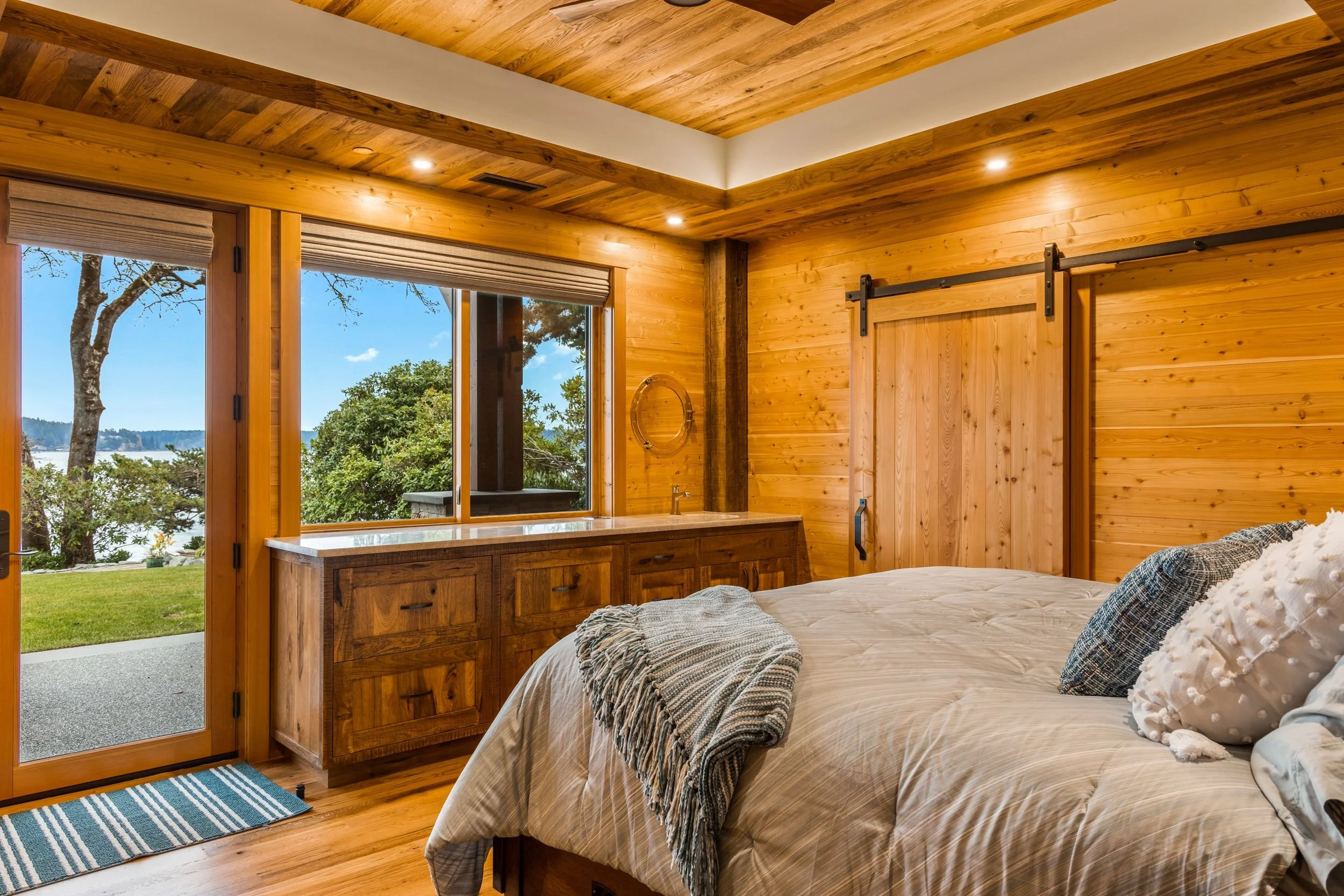 A cozy bedroom with wooden walls, a large bed with pillows and a blanket, a wooden sideboard below windows showing a view of trees and water outside, and a sliding barn door.