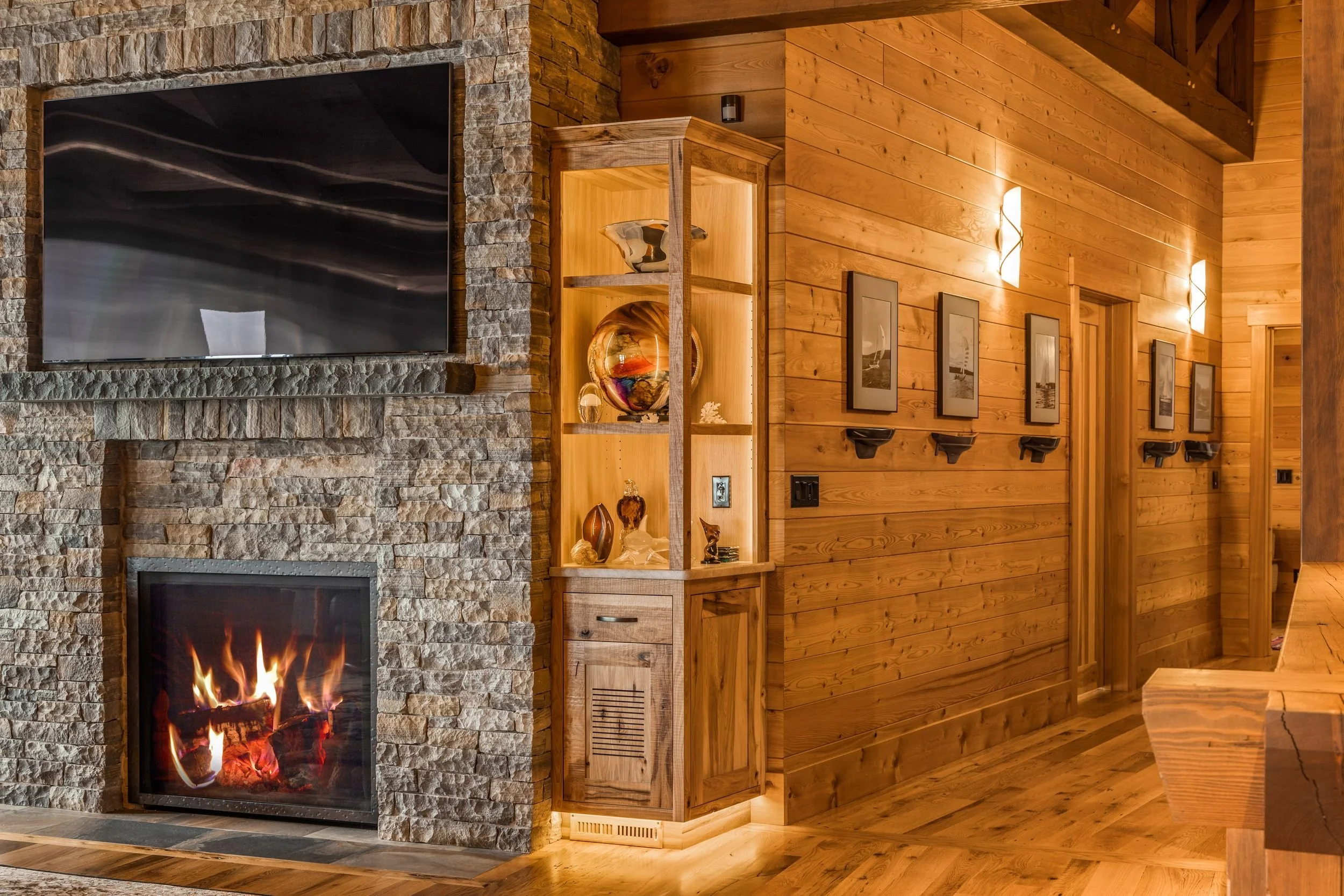 Living room with stone fireplace, mounted flat-screen TV, wooden wall panels, a built-in wooden display cabinet with decorative glass art, framed pictures with shelf lights on the wall, warm wooden flooring, and a staircase in the background.