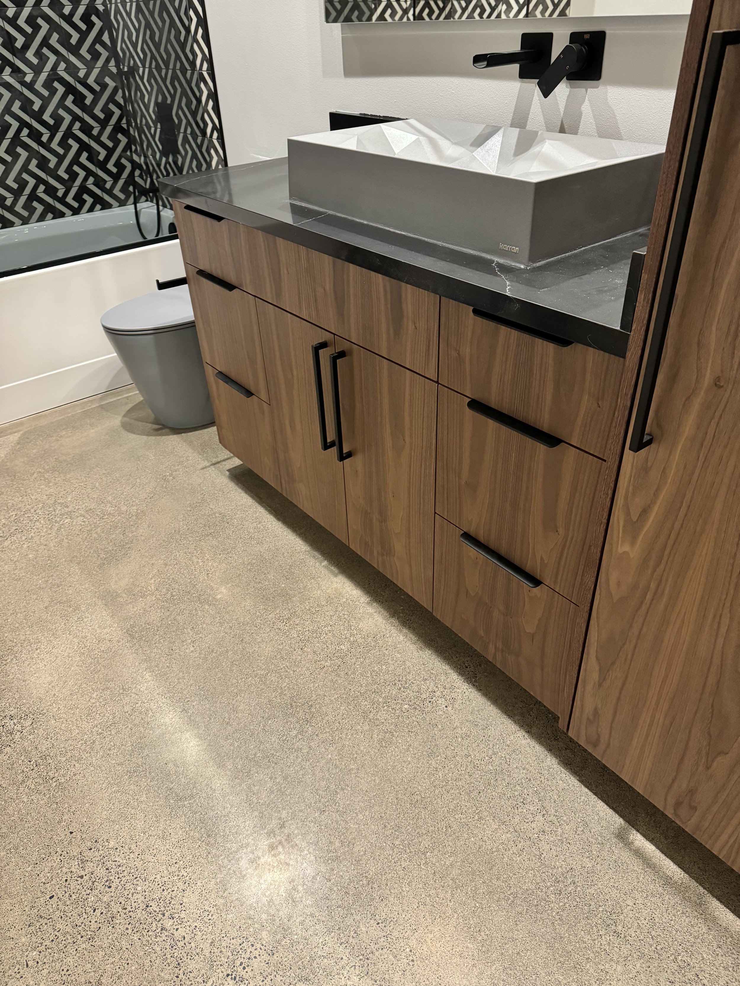 Modern bathroom vanity with a black countertop and a geometric, angular white vessel sink. Wooden cabinet with black handles, a gray toilet, and patterned black and white shower surrounded by white walls.
