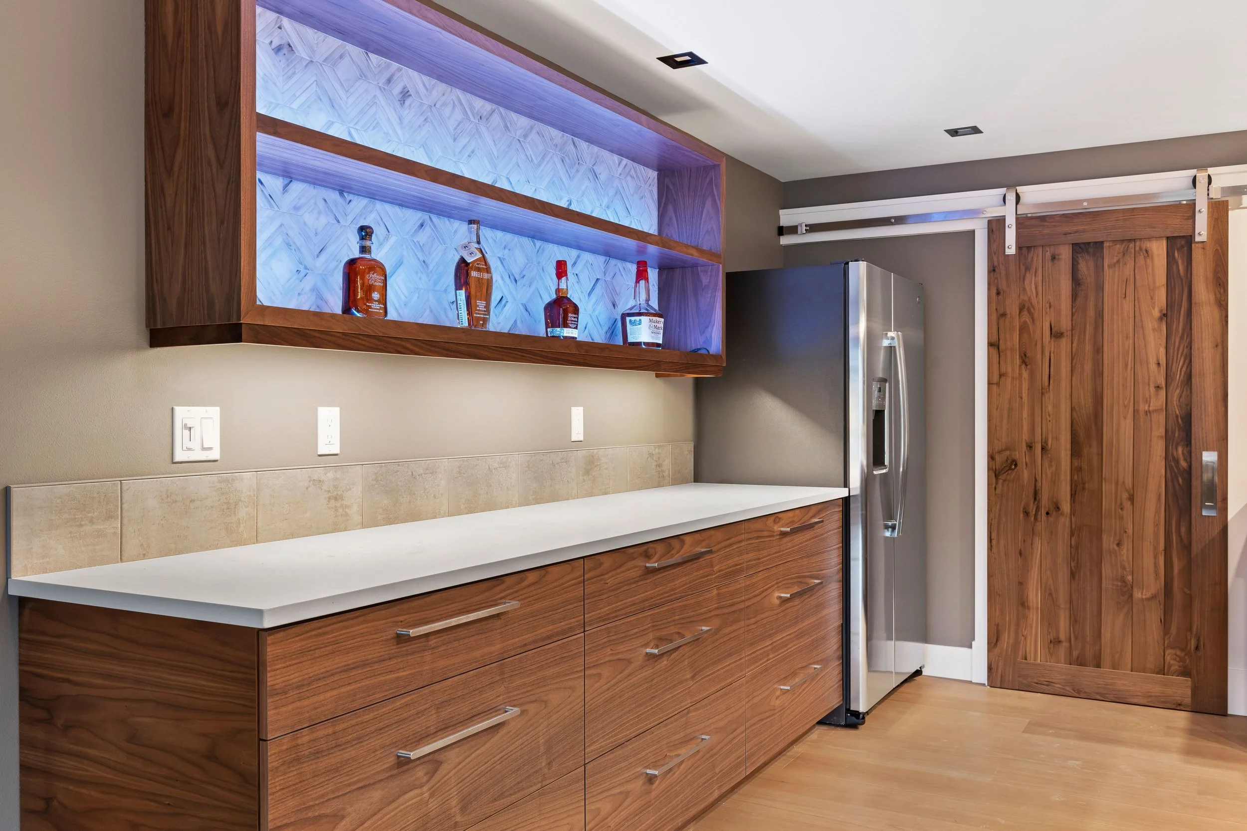 Modern kitchen with wooden cabinets, a white countertop, and a stainless steel refrigerator. A wooden sliding barn door and a wall-mounted liquor shelf with bottles are visible.