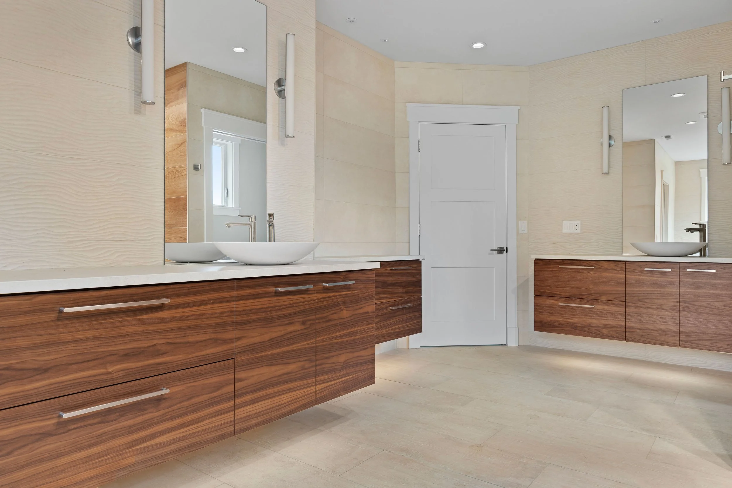 Modern bathroom with two vessel sinks, wooden cabinetry, large mirrors, and light-colored walls and flooring.