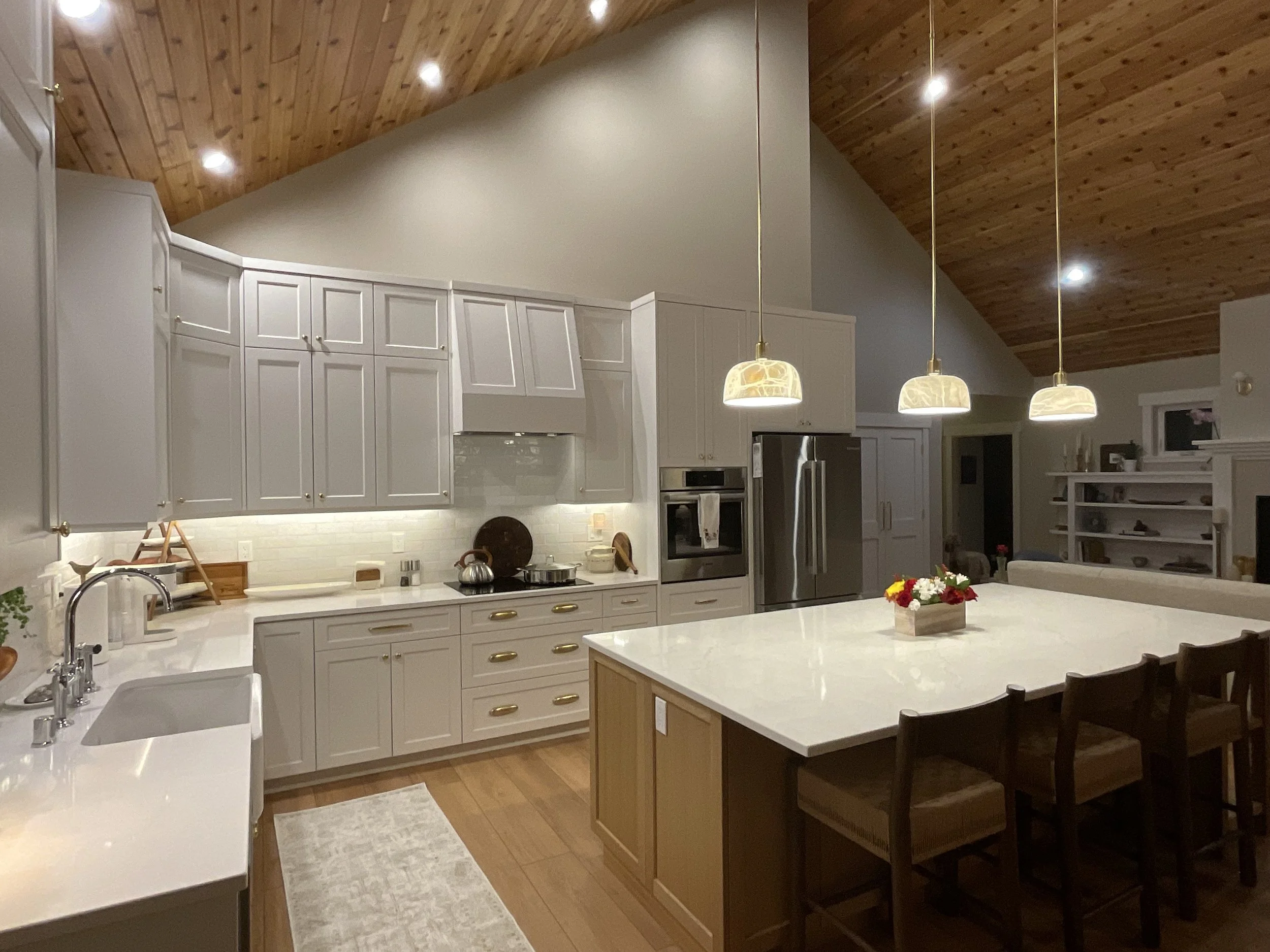 A cozy kitchen with white cabinets, a white countertop, and a large island with seating. The kitchen features a stainless steel refrigerator and oven, wooden ceiling with pendant lights, and a small rug in front of the sink.
