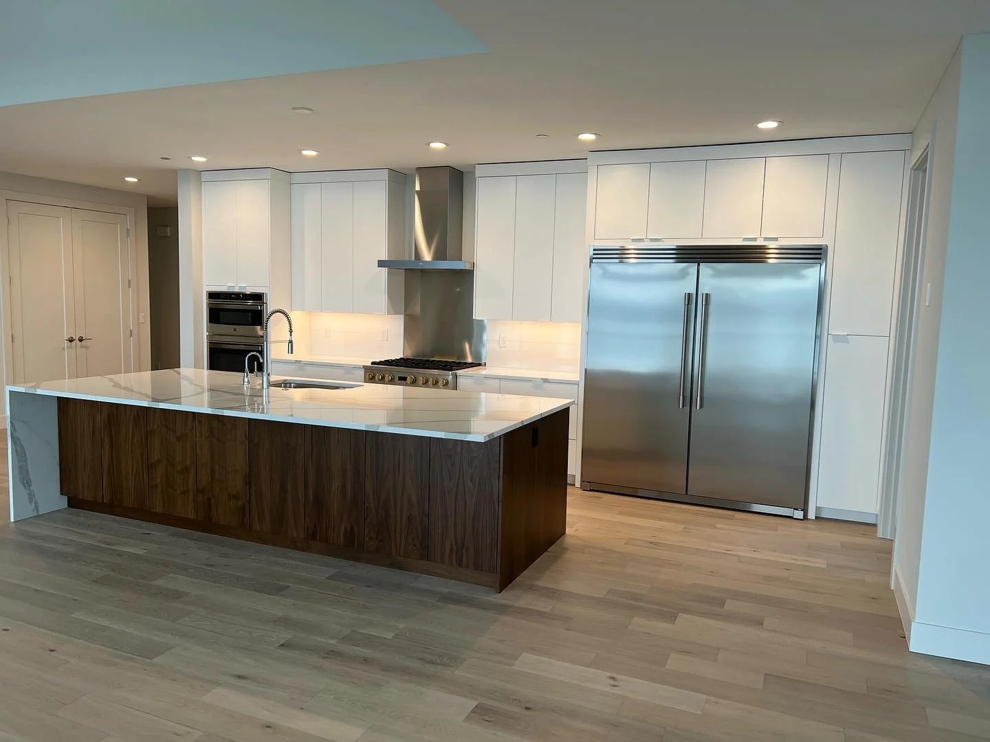 Modern kitchen with white cabinets, a large wooden kitchen island with a white countertop, stainless steel appliances including a refrigerator, oven, and stove with a range hood, and hardwood floors.