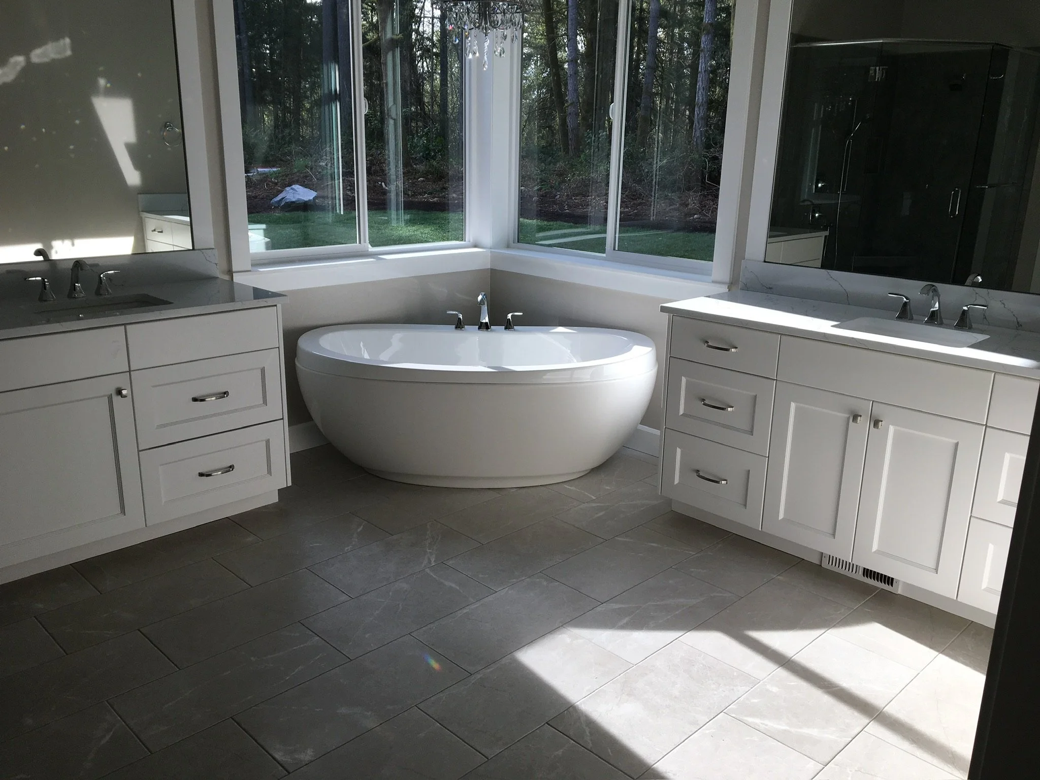 Modern bathroom with a freestanding bathtub, two white vanities with sinks, large windows showing a green outdoor area, and a mirror on the right wall.