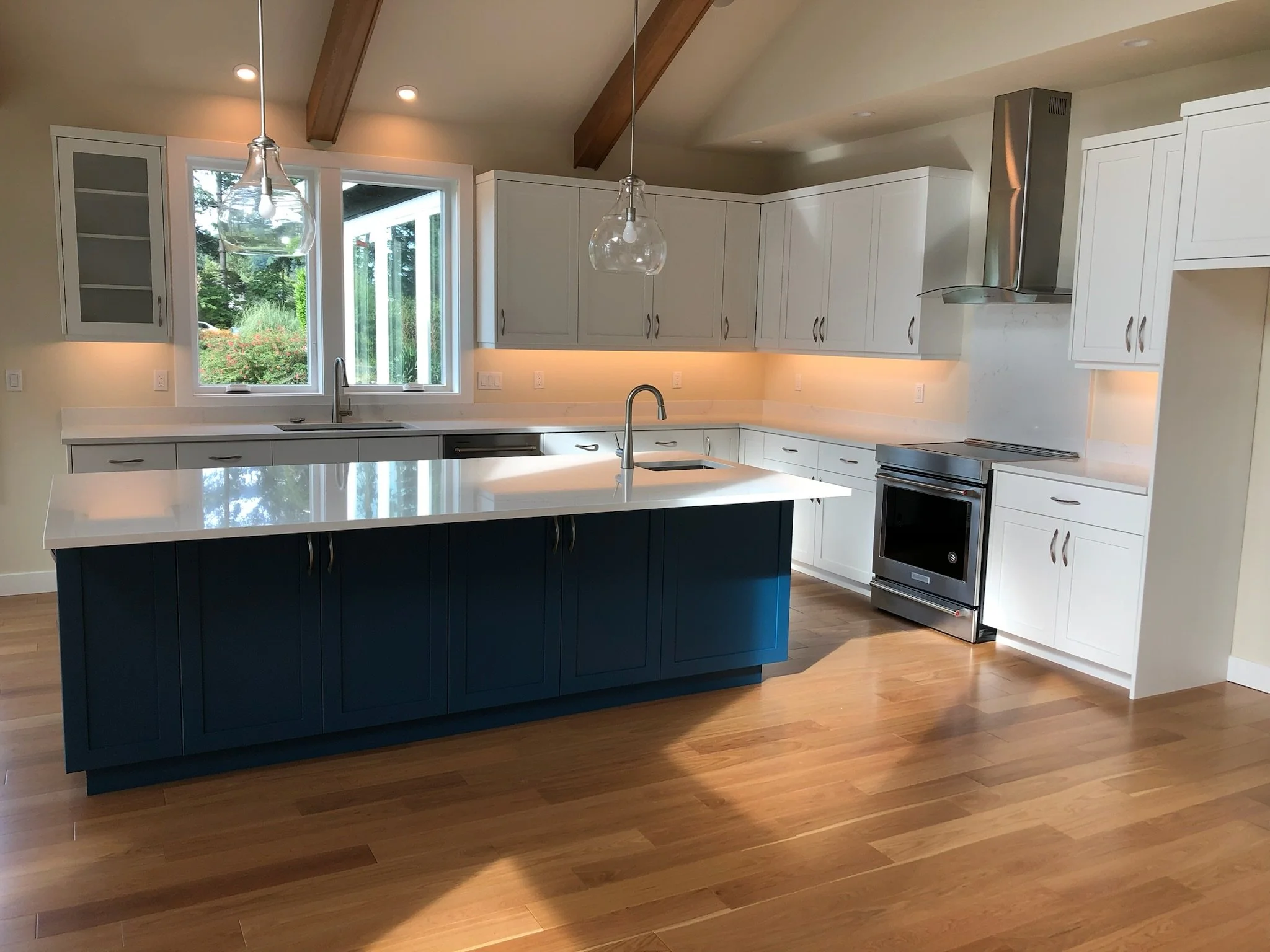 Modern kitchen with white cabinets, a blue island with a white countertop, hardwood flooring, and stainless steel appliances including a stove and range hood. There are pendant lights and a window above the sink.