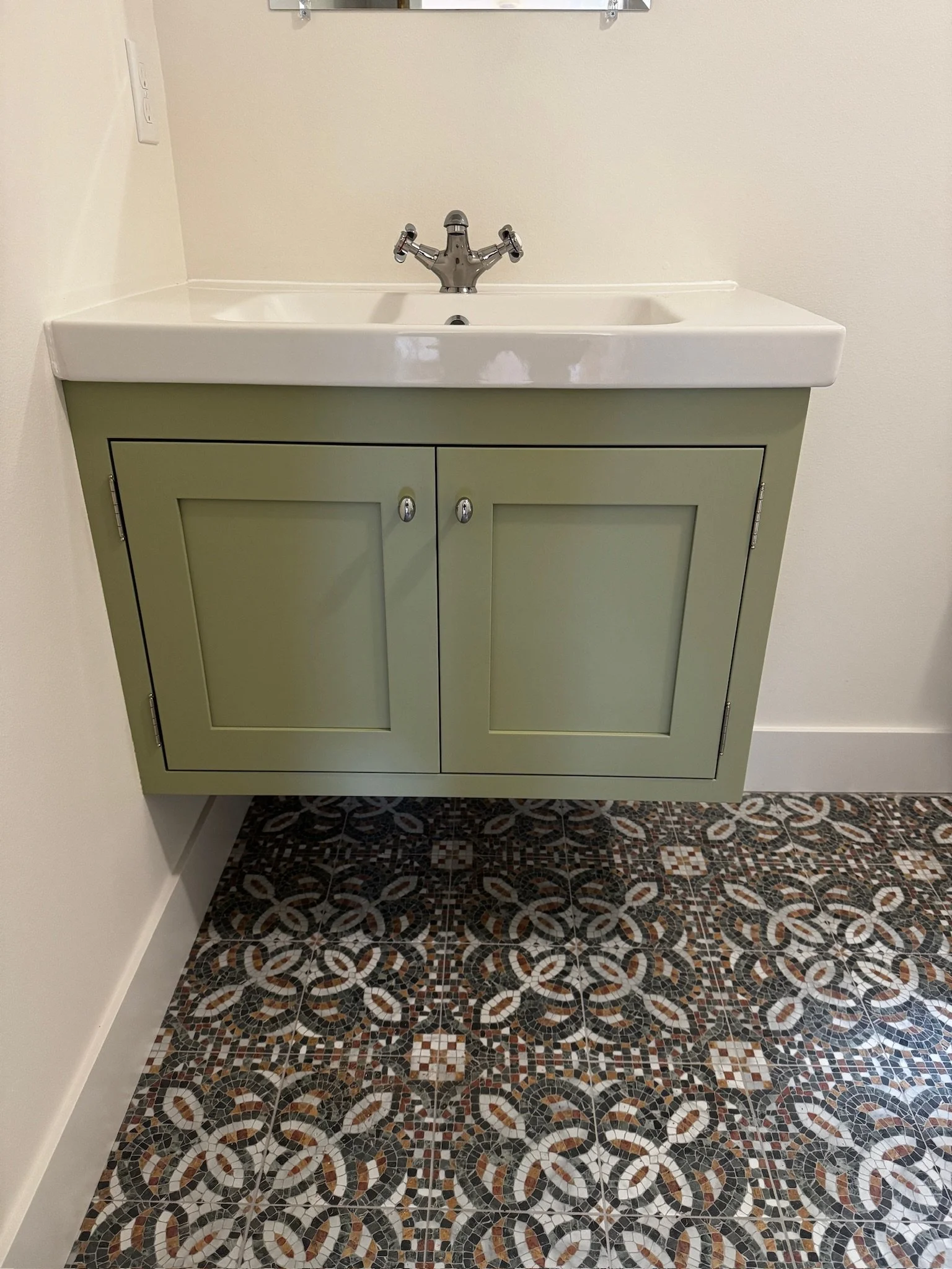 A bathroom vanity with a white ceramic sink and a green cabinet below, set against a plain wall. The floor has a patterned mosaic tile design in shades of black, white, tan, and gray.