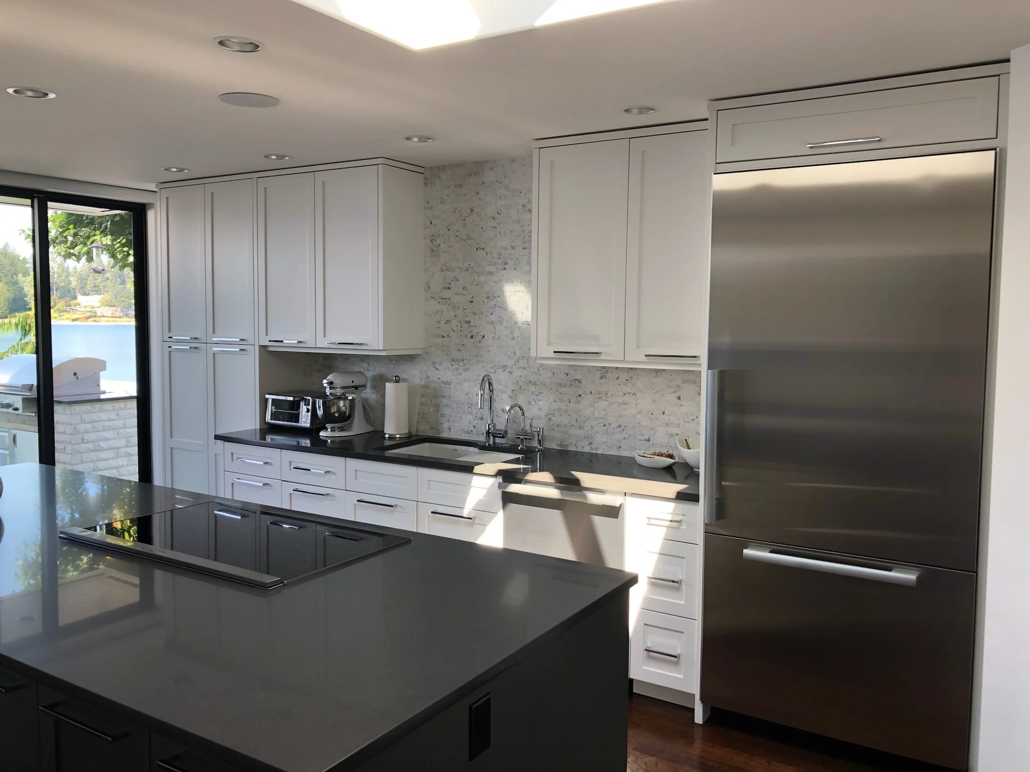 Modern kitchen with white cabinets, black countertops, stainless steel refrigerator, and a view of a lake outside through sliding glass doors.