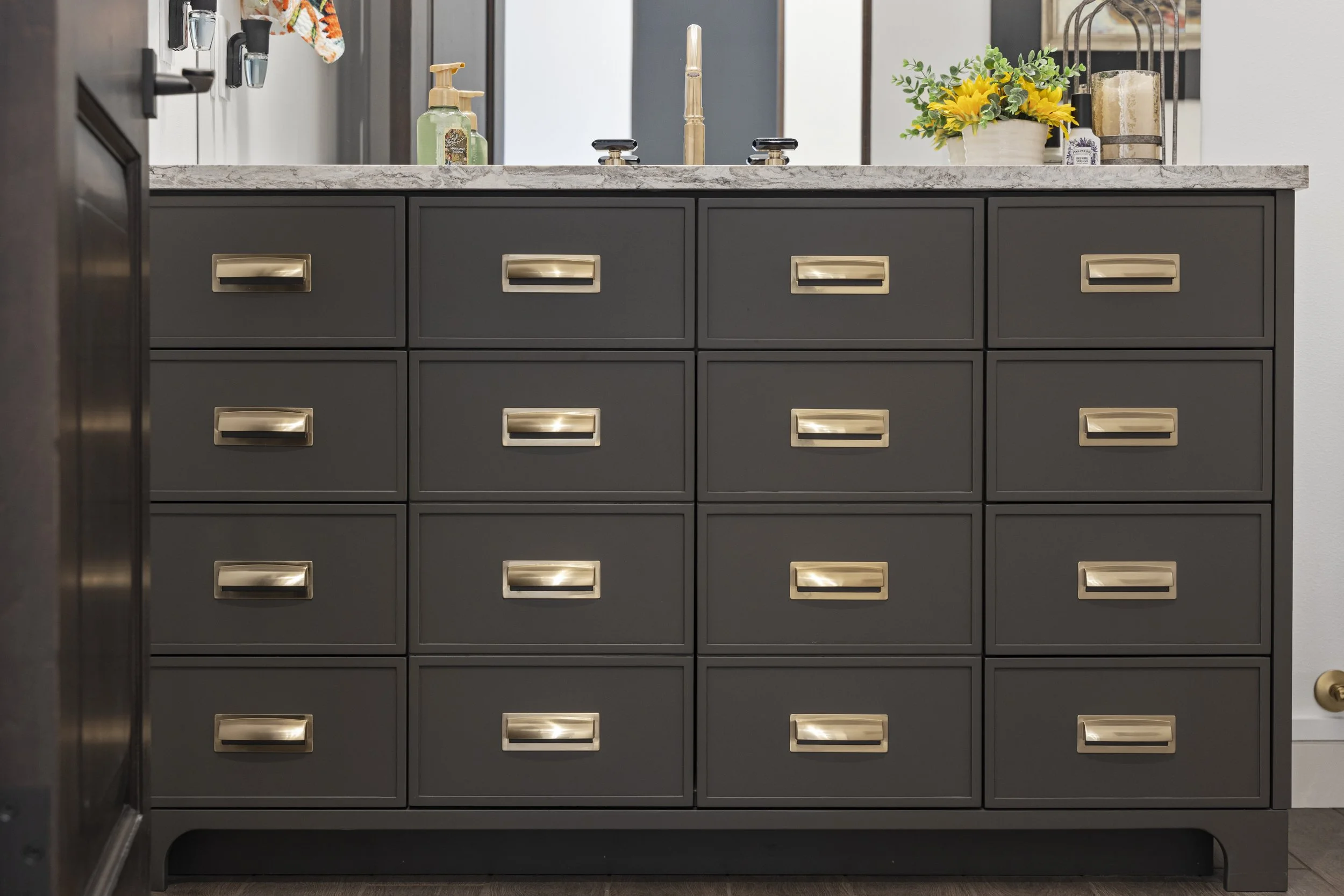 Gray dresser with 15 drawers, each with a gold handle, topped with a white marble countertop. On top are a green soap dispenser, a small bottle, a sunflower arrangement in a white pot, a lantern with a candle, and a soap pump. A mirror with a black f