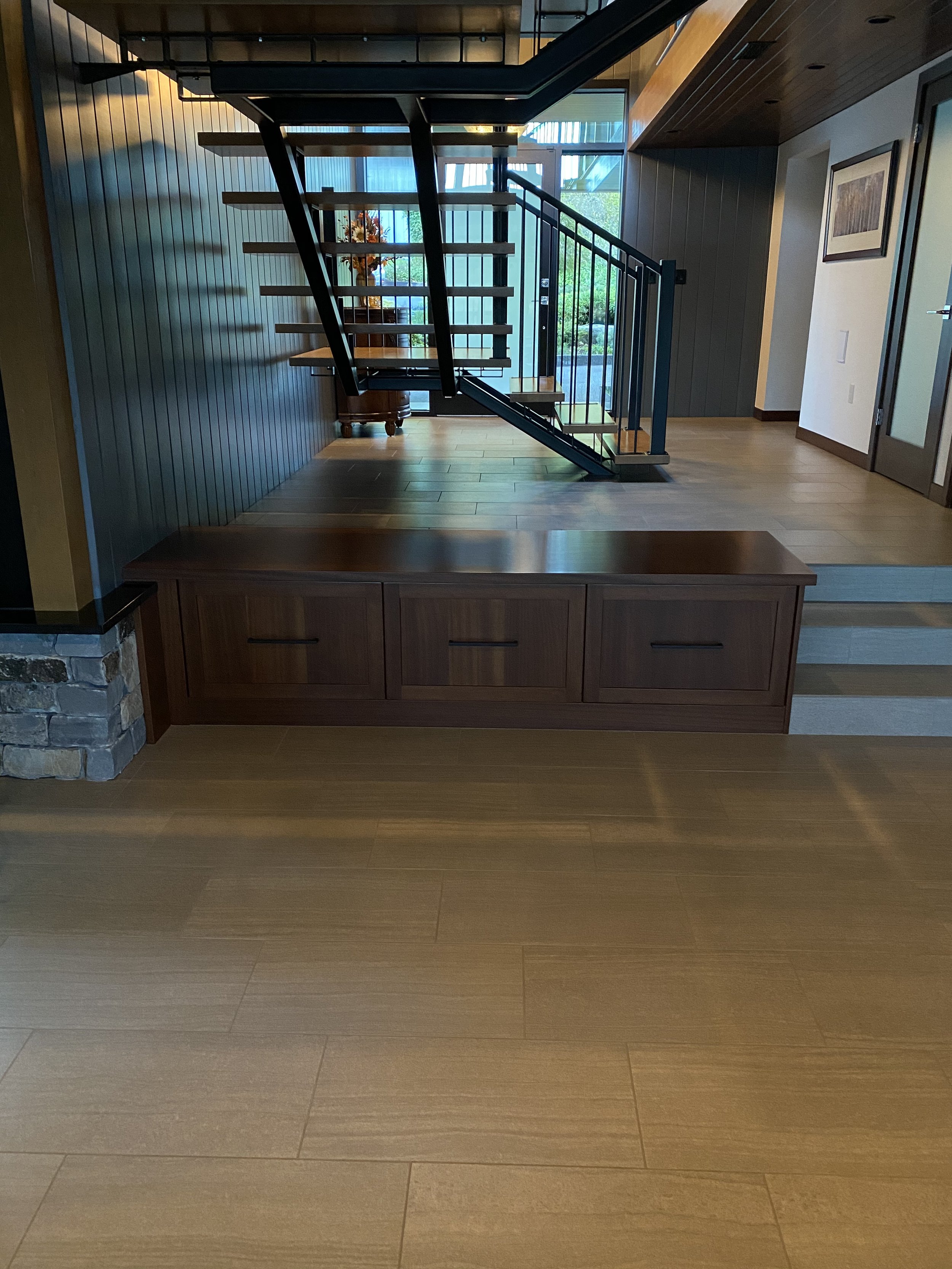 Interior view of a modern home with wooden flooring, a staircase with black metal railings, a side table with flowers, and large windows leading outside.