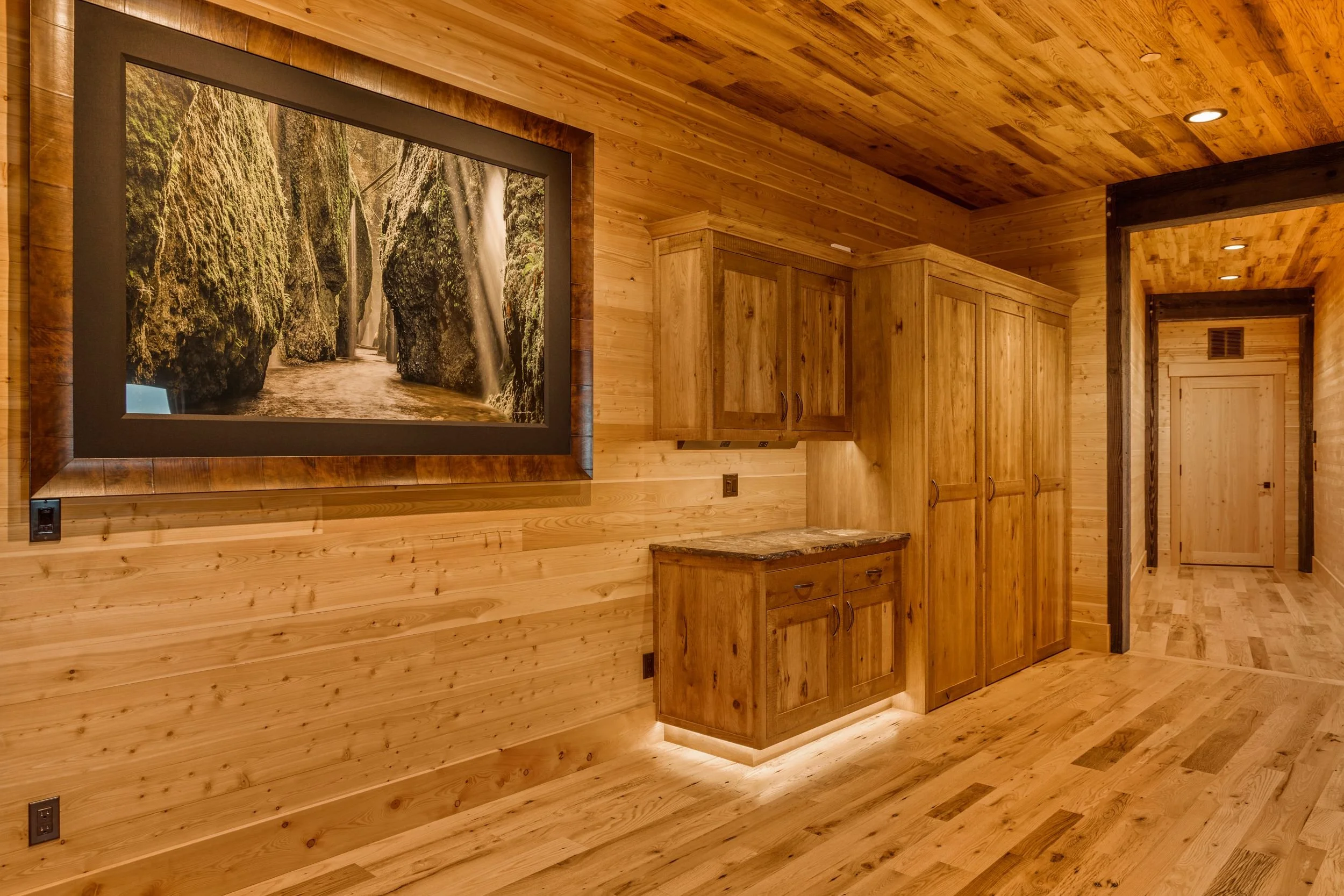 Interior view of a wooden room with wood-paneled walls and ceiling, featuring a large framed nature photograph of a river and greenery, and wooden cabinetry.