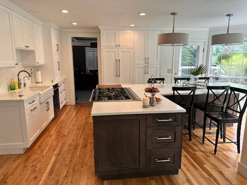 Modern kitchen with white cabinetry, a dark island, wooden floors, and a dining area with black chairs and a table near large windows.