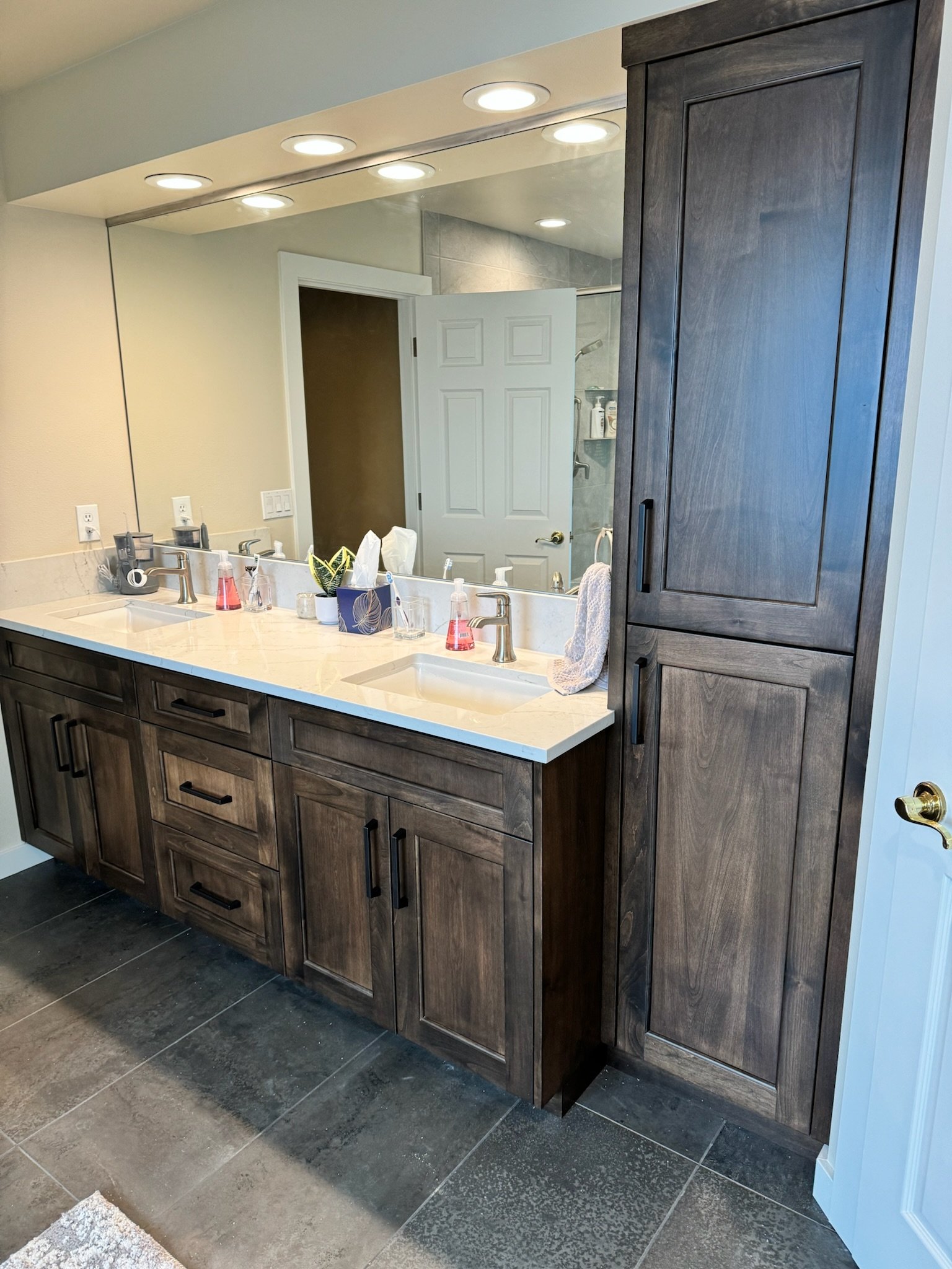 A double vanity bathroom with two sinks, a large mirror, dark wooden cabinets, and a wall cabinet. The countertop has soap dispensers, a tissue box, and towels. In the background, a door and a walk-in shower are visible.
