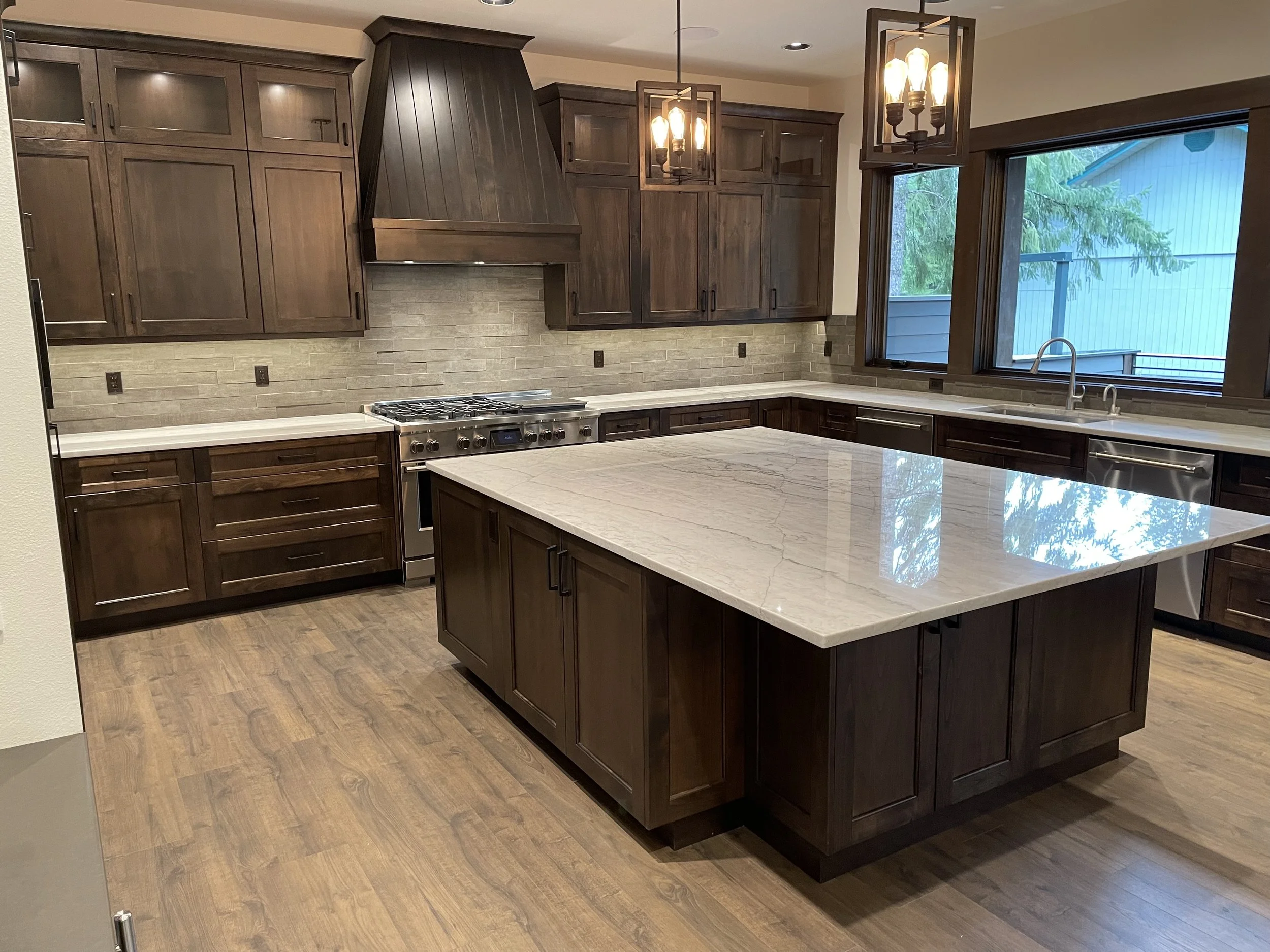 A modern kitchen with dark wooden cabinets, beige stone backsplash, a large white marble island, stainless steel appliances, and two pendant lights hanging above the island. There are large windows overlooking greenery.