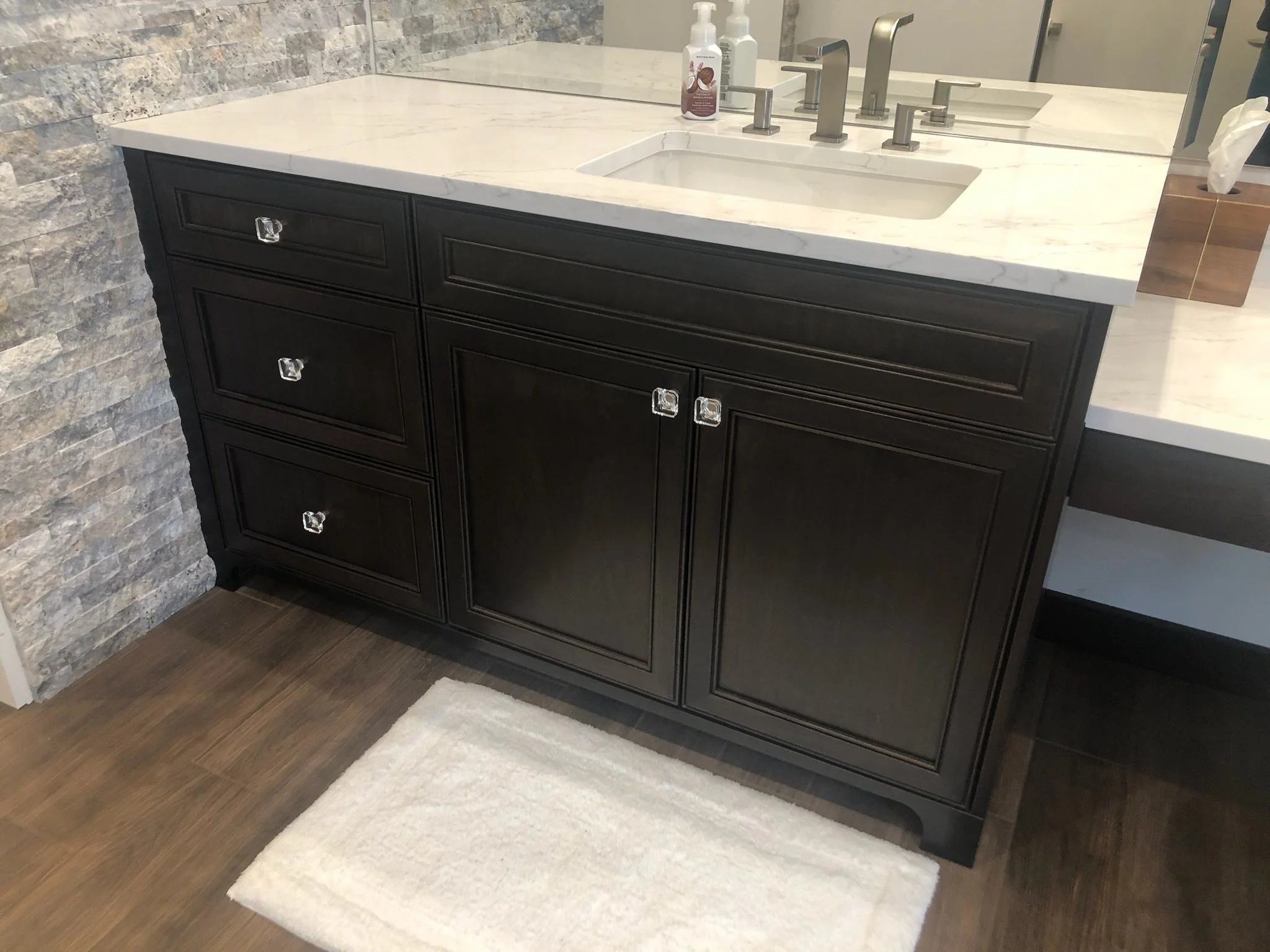 Bathroom vanity with a white marble countertop, black wooden cabinets, a built-in sink, a silver faucet, a soap dispenser, a tissue box, a mirror, and a textured stone wall.