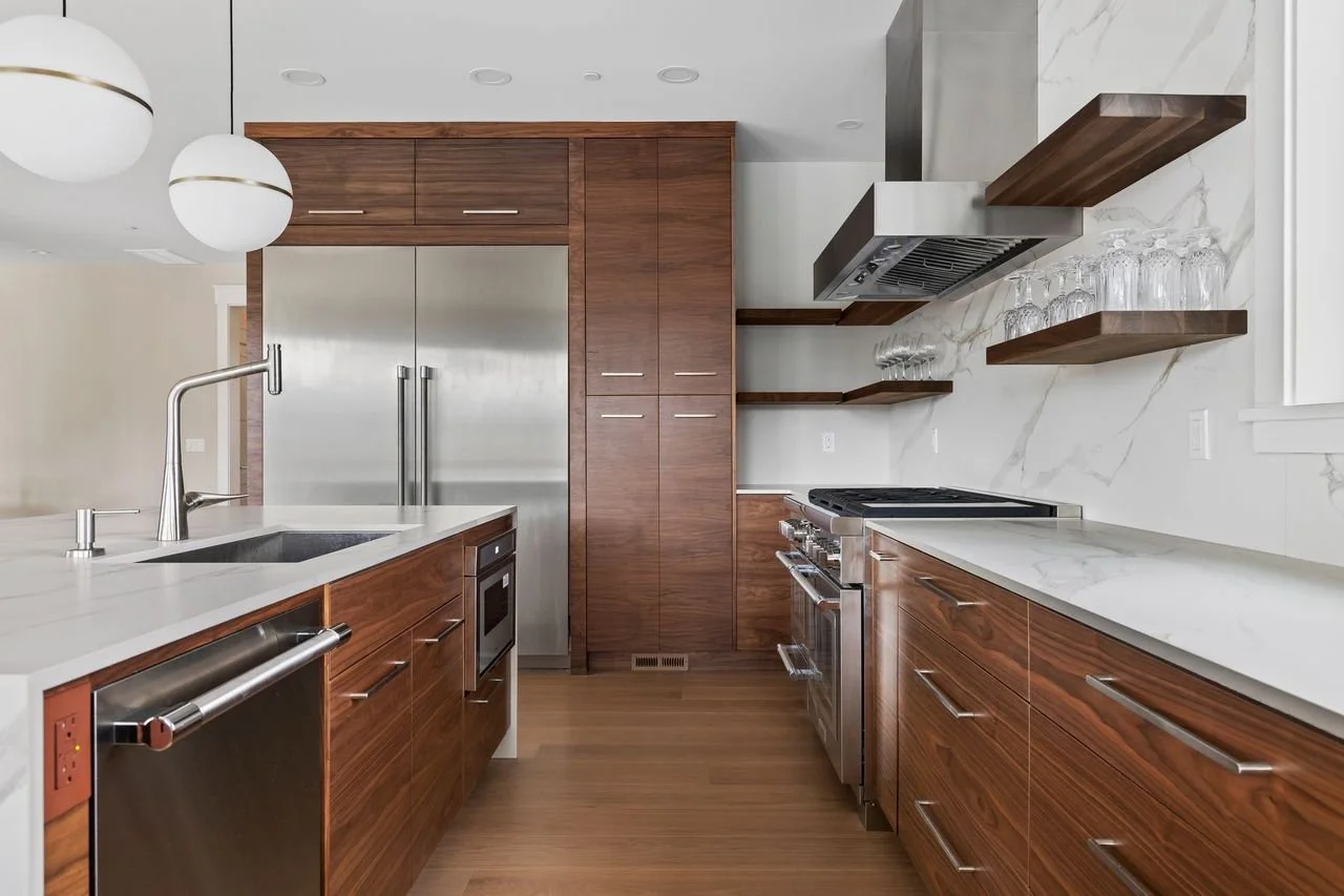 Modern kitchen with wooden cabinets, white marble countertops, stainless steel appliances, and open wooden shelves with glassware. Recessed lighting and two white spherical pendant lights hang above the island.