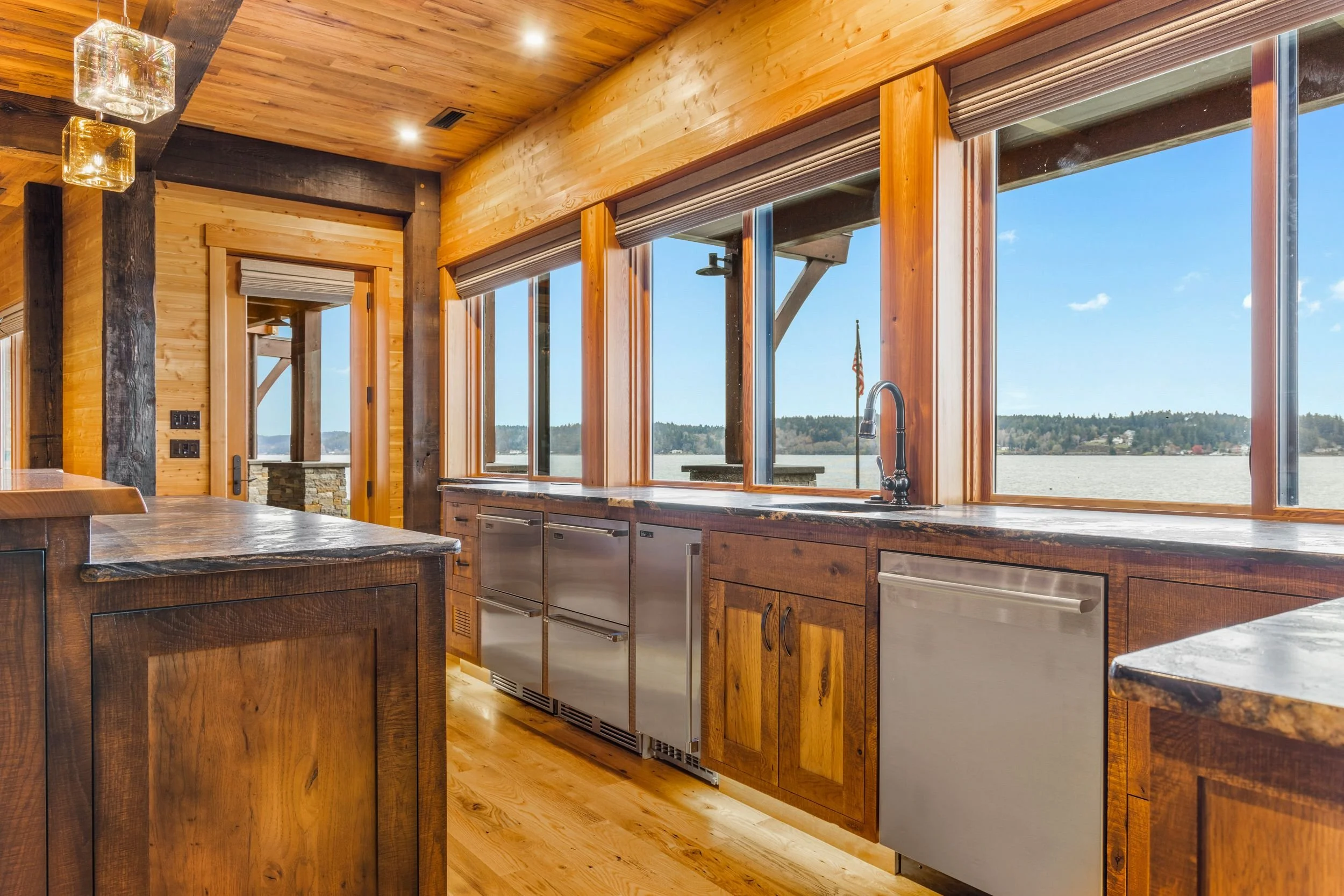 Interior view of a rustic kitchen with wooden cabinets, large windows overlooking a body of water, and stainless steel appliances.