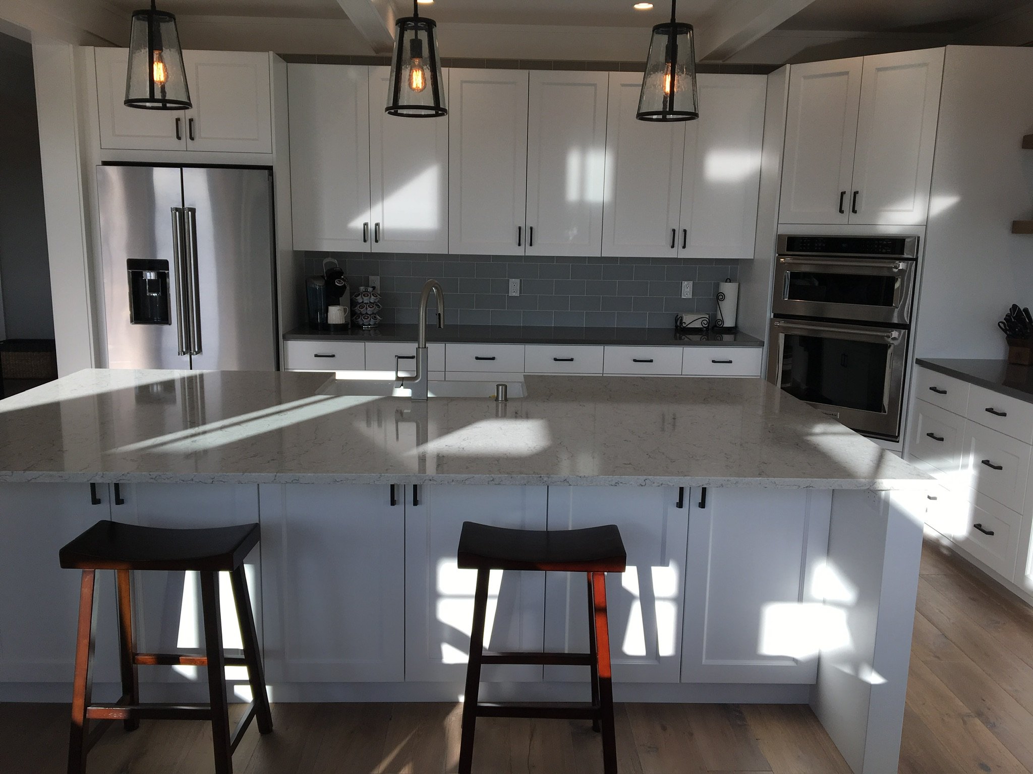 Modern kitchen with white cabinets, gray backsplash, stainless steel refrigerator, double ovens, and a large marble island with two wooden bar stools.