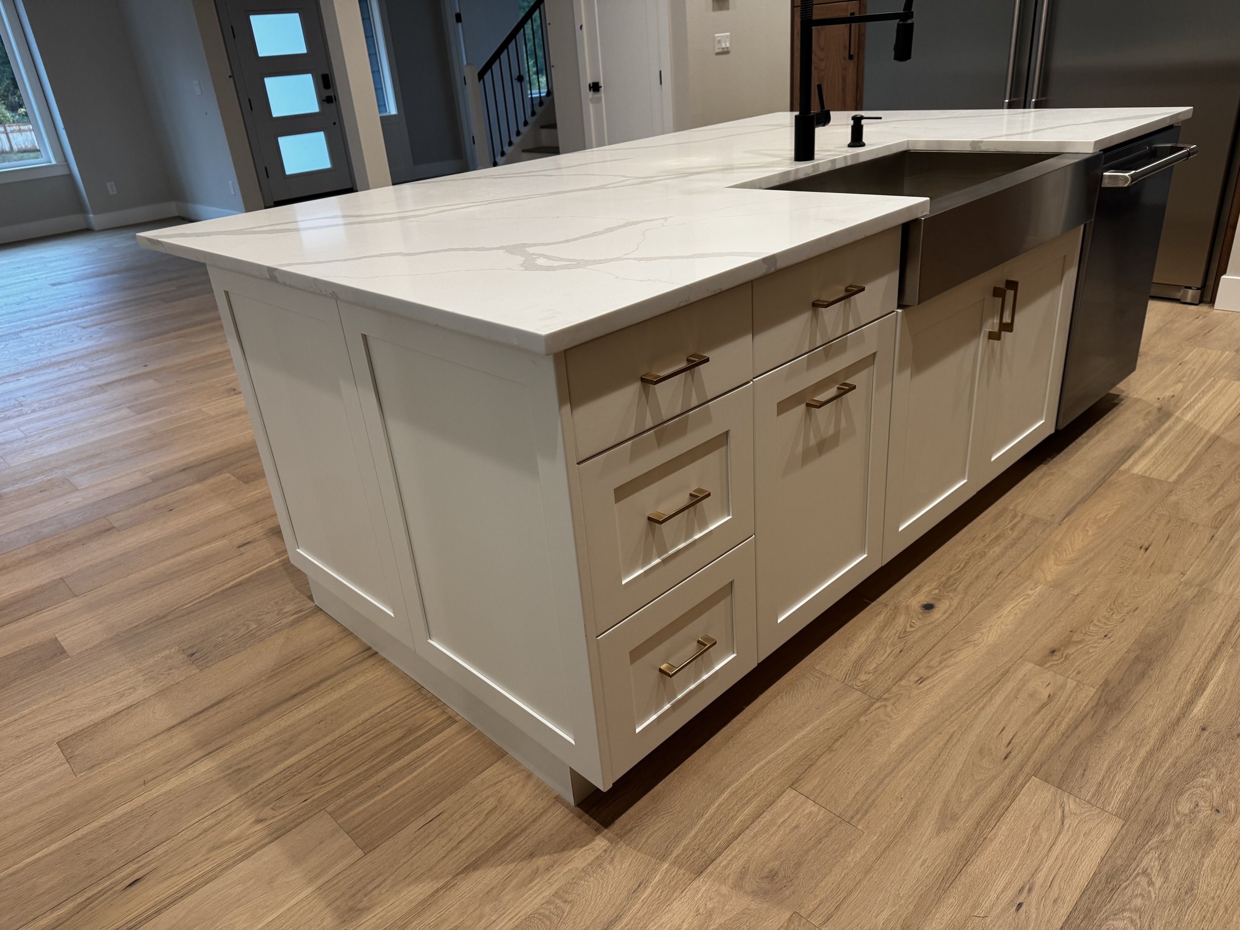 Kitchen island with white marble countertop, cream-colored cabinet base with drawers and cabinets, black farmhouse sink, black faucet, and hardwood flooring.