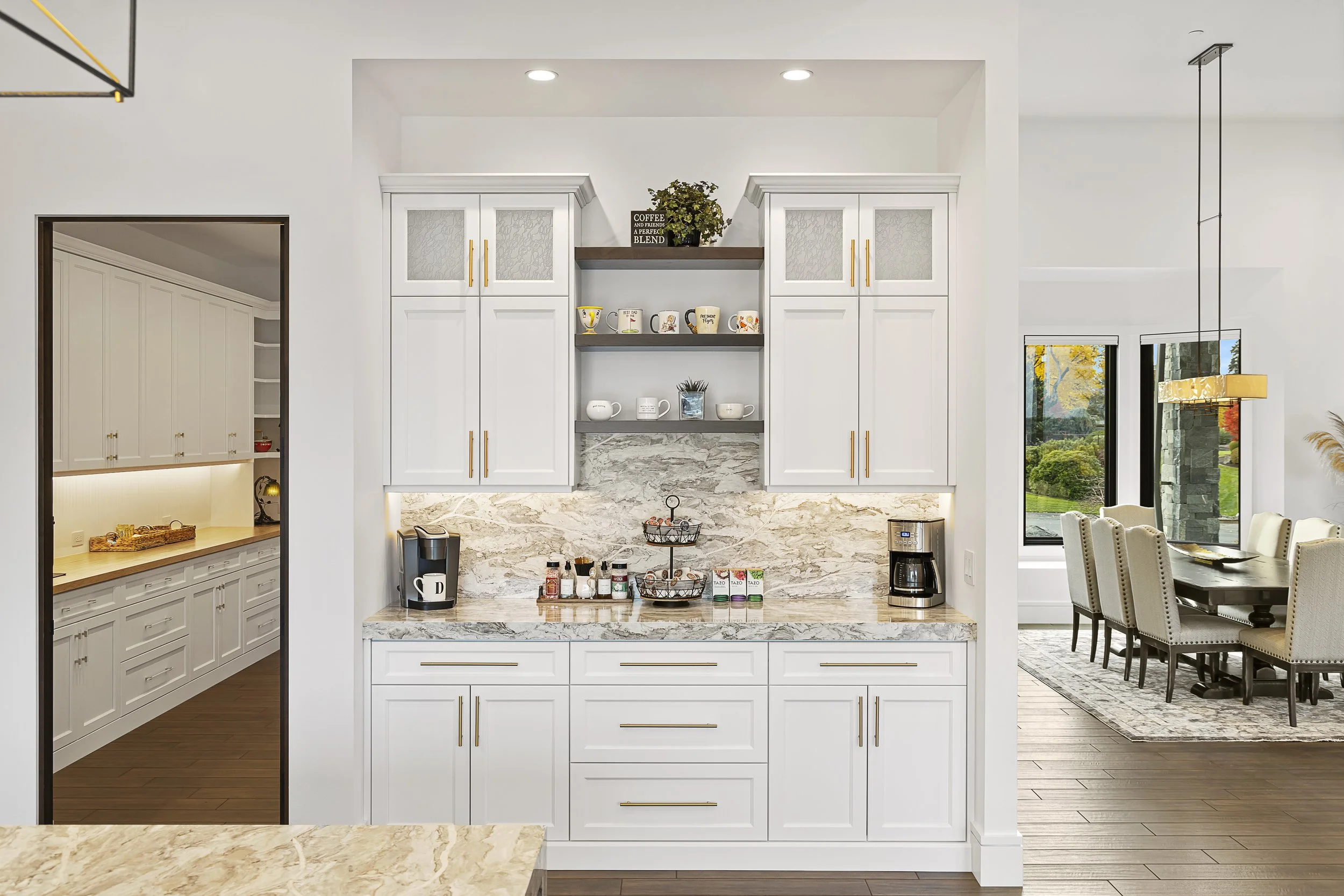 Modern kitchen with white cabinets, marble countertops, open shelving with coffee mugs and decor, coffee maker, and a view of a dining area with large windows showing greenery outside.