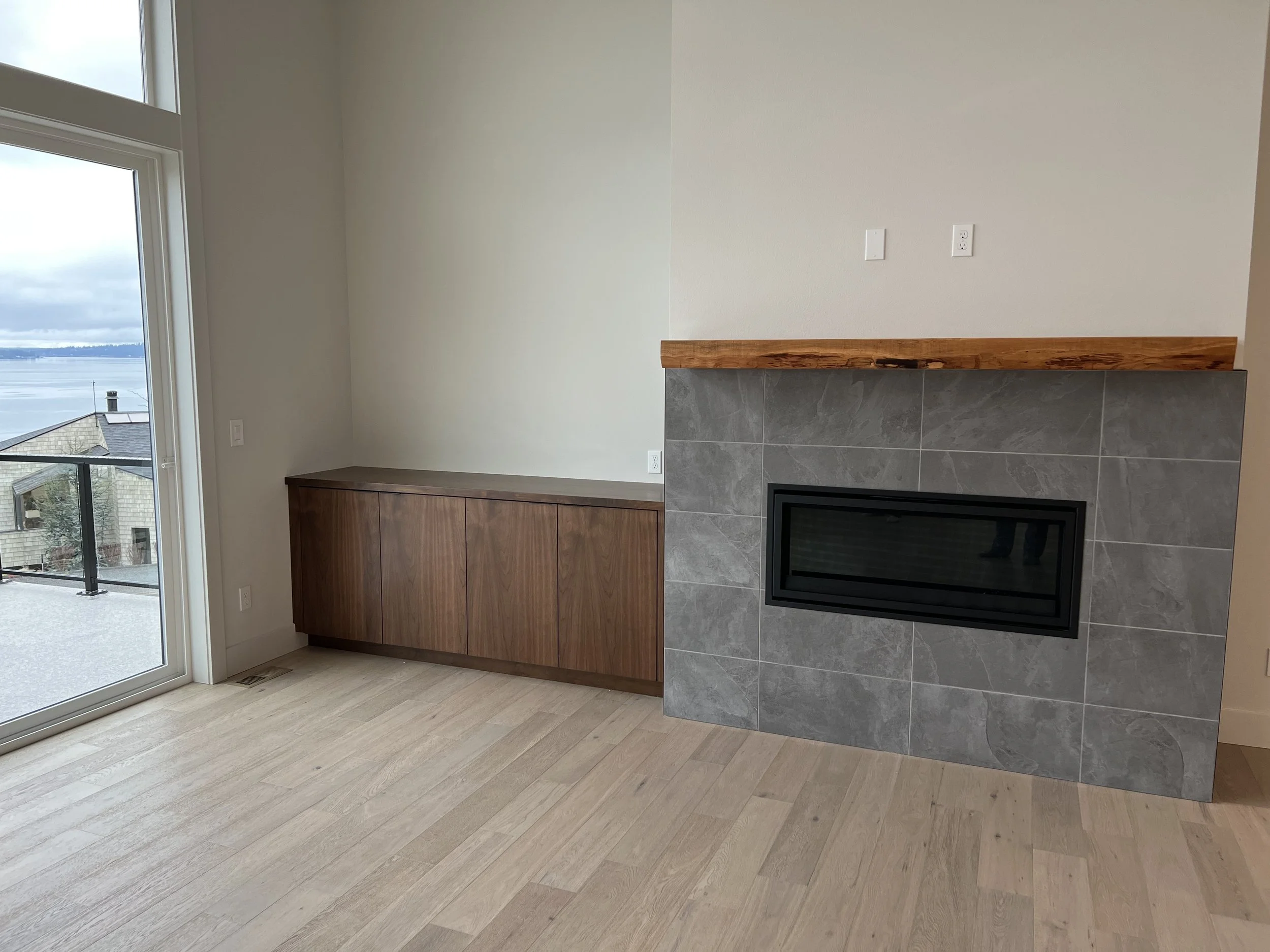 Modern living room with a fireplace, gray tile surround, wooden mantle, and a built-in wooden cabinet, next to a glass door leading to a balcony with a view of water and cloudy sky.