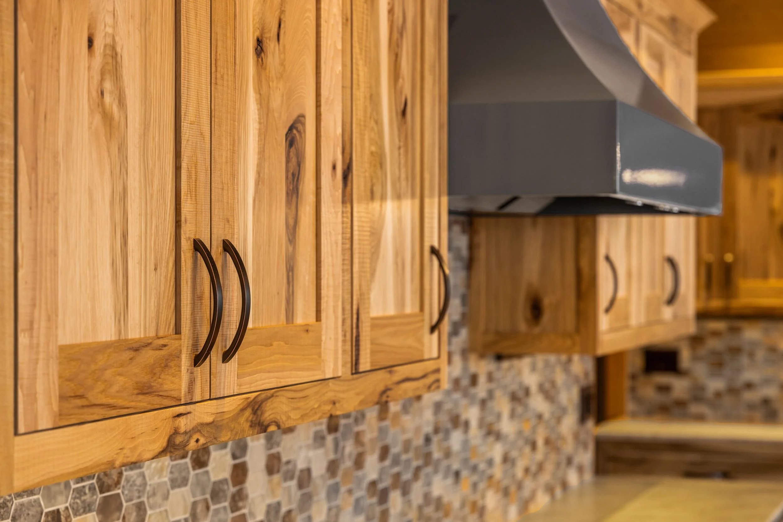Kitchen with wooden cabinets, black handles, and a gray range hood above a mosaic tile backsplash.