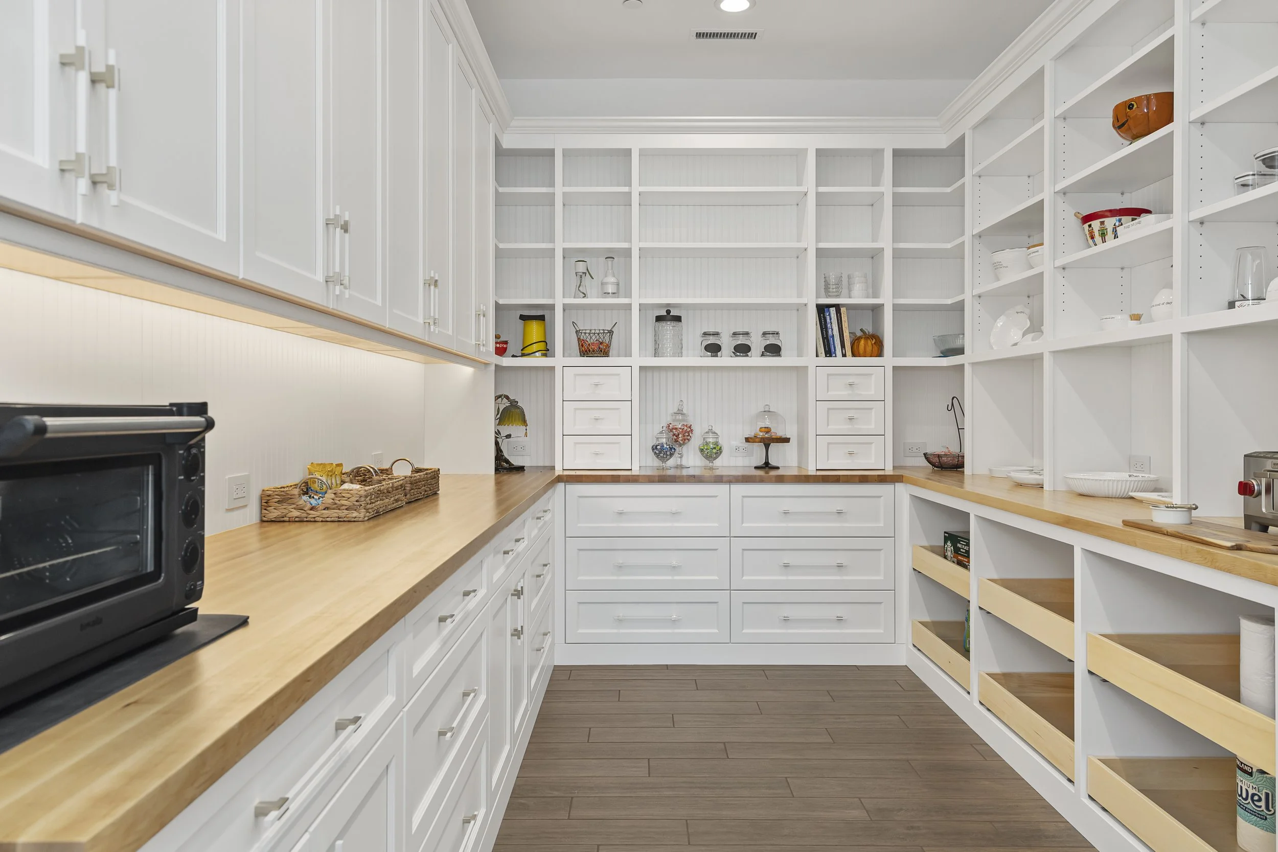 Interior view of a spacious walk-in pantry with white cabinetry and open shelves, wooden countertops, and a wooden floor. The shelves are partially stocked with glass jars, bowls, and decorative items, with some empty shelf space available.