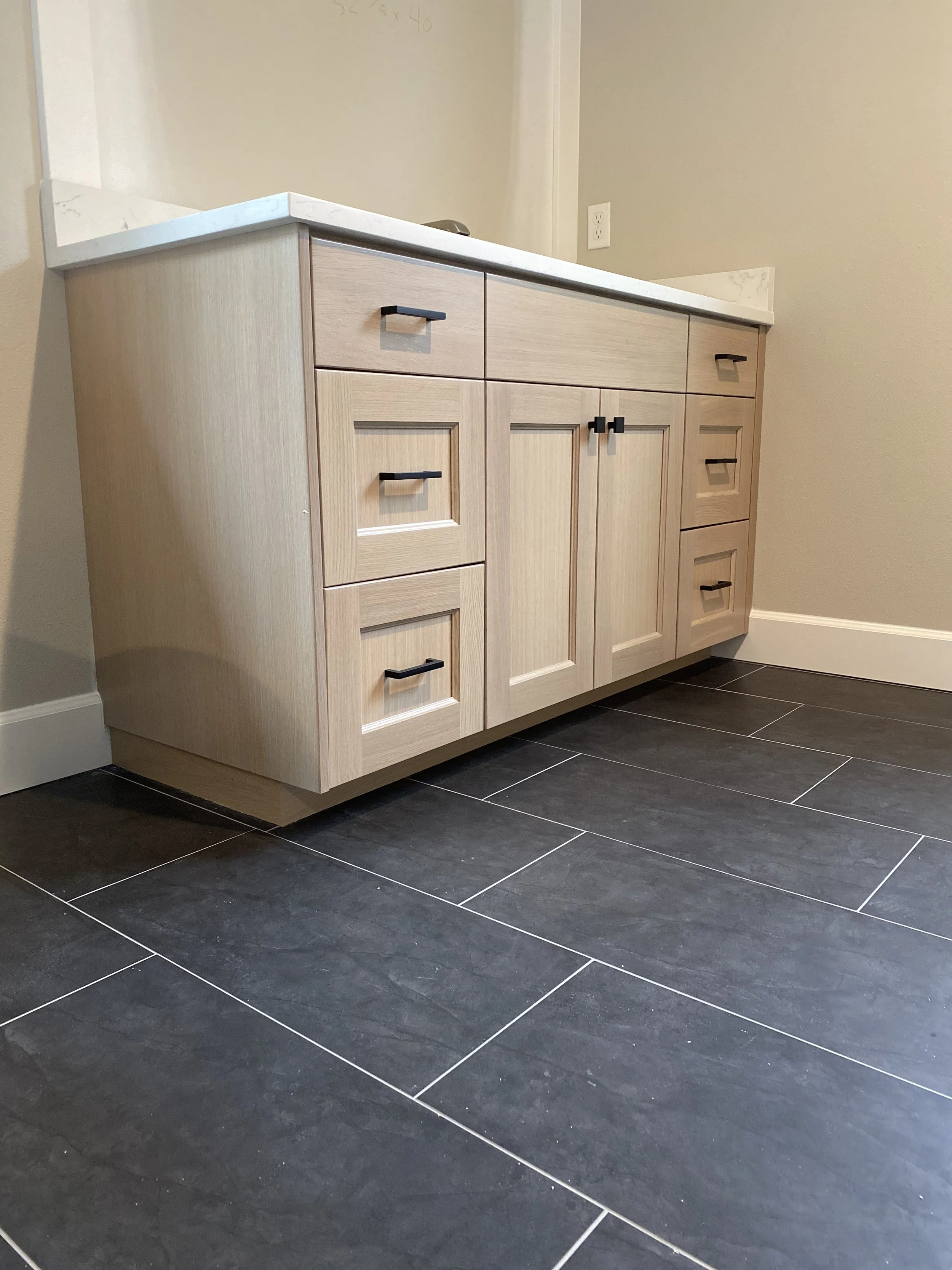 Light wood bathroom vanity with black handles, marble countertop, and black floor tiles.