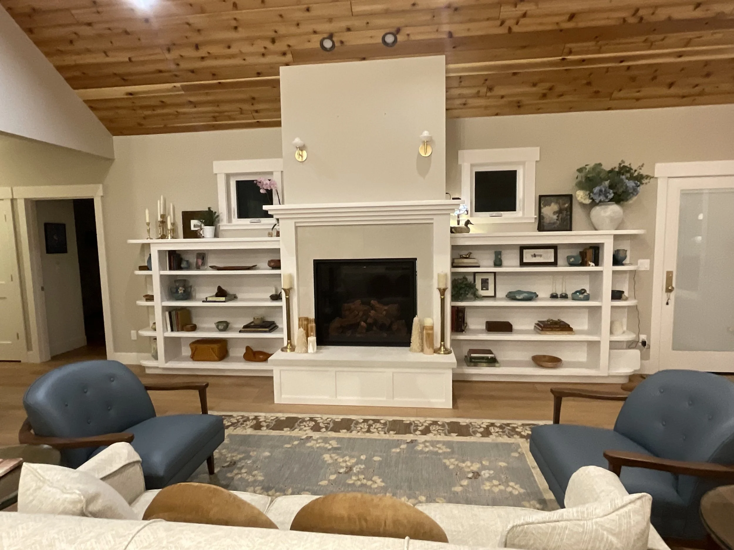A living room with a fireplace, built-in white shelves, and wood-paneled ceiling. Two blue armchairs and a couch are visible, along with decorative items and books on the shelves.