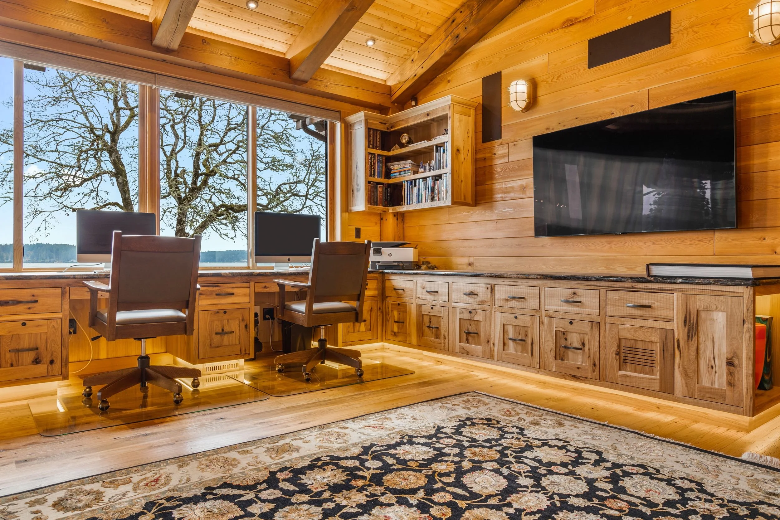 Home office with large window showing tree and waterfront, wooden walls and ceiling, built-in desk with two chairs, bookshelf, flat-screen TV, and an area rug.