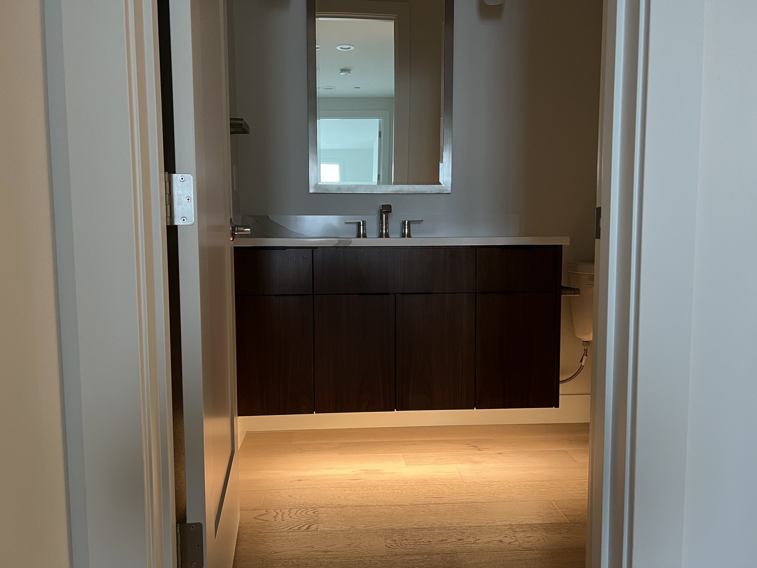 View through a doorway into a modern bathroom with a dark wooden vanity, double sink, large mirror, and window.