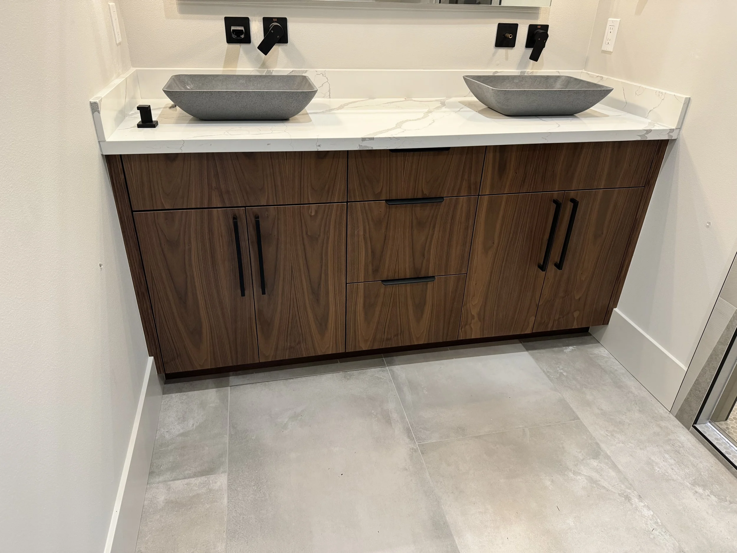 Wooden vanity with two gray stone vessel sinks on a white marble countertop in a bathroom.