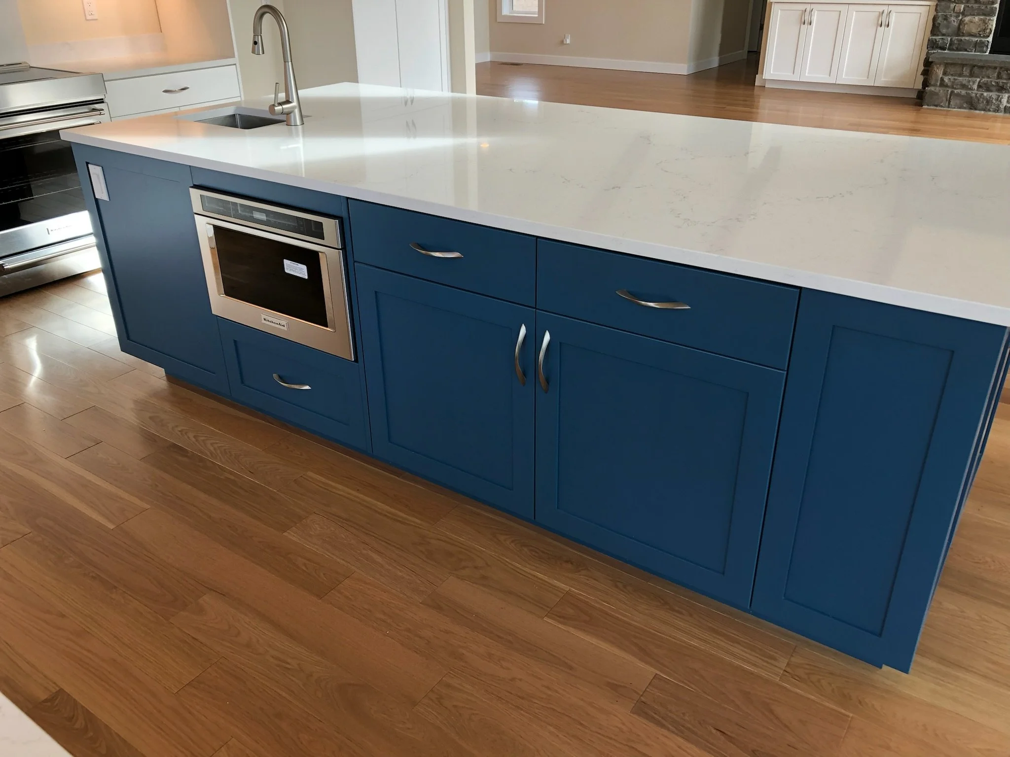 A kitchen island with a blue base and a white countertop. The island has storage cabinets and drawers, a built-in microwave, a sink with a faucet, and wooden flooring.
