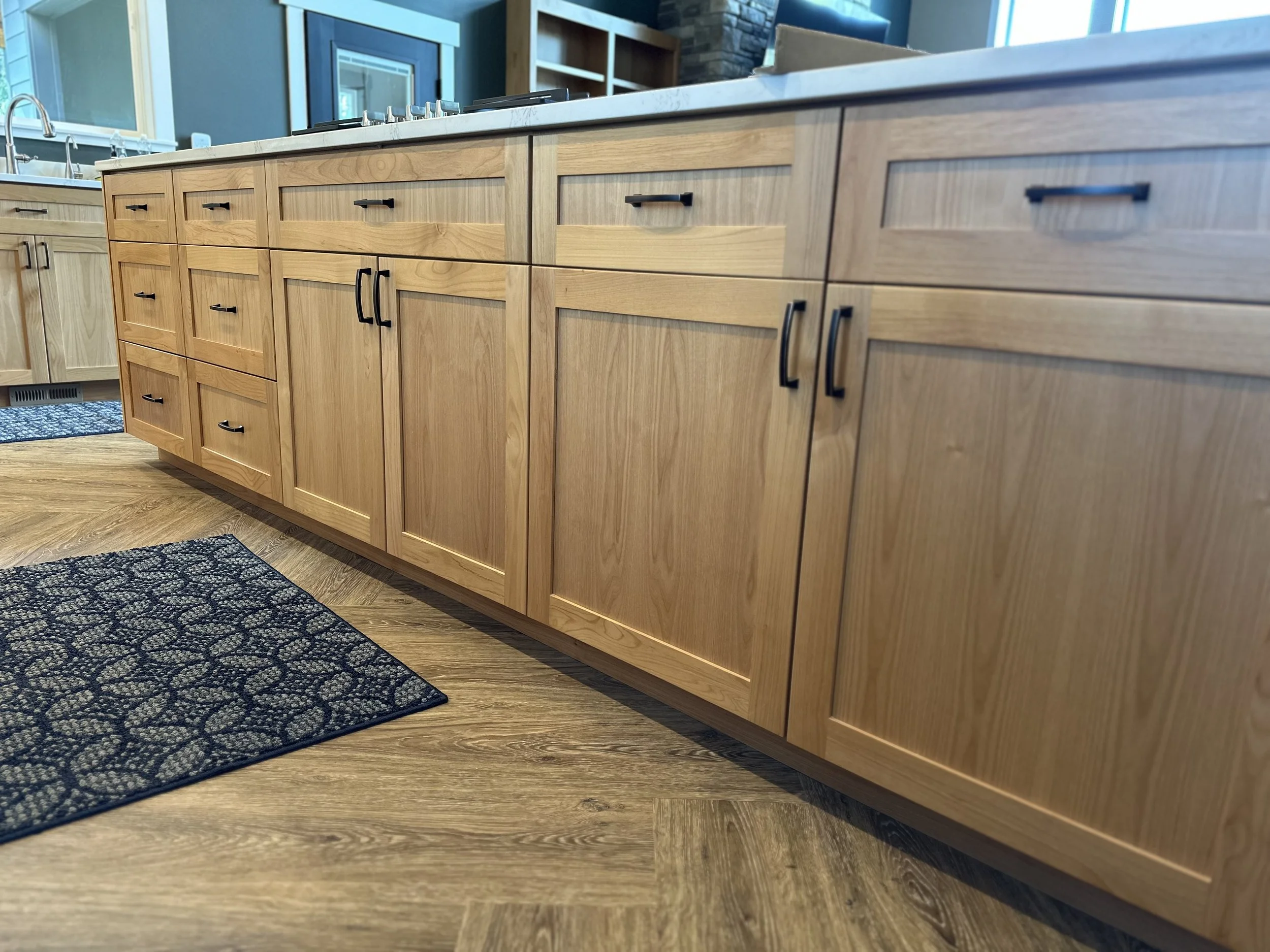 Wooden kitchen cabinets with black handles, a white countertop, and a black patterned rug on wood flooring.