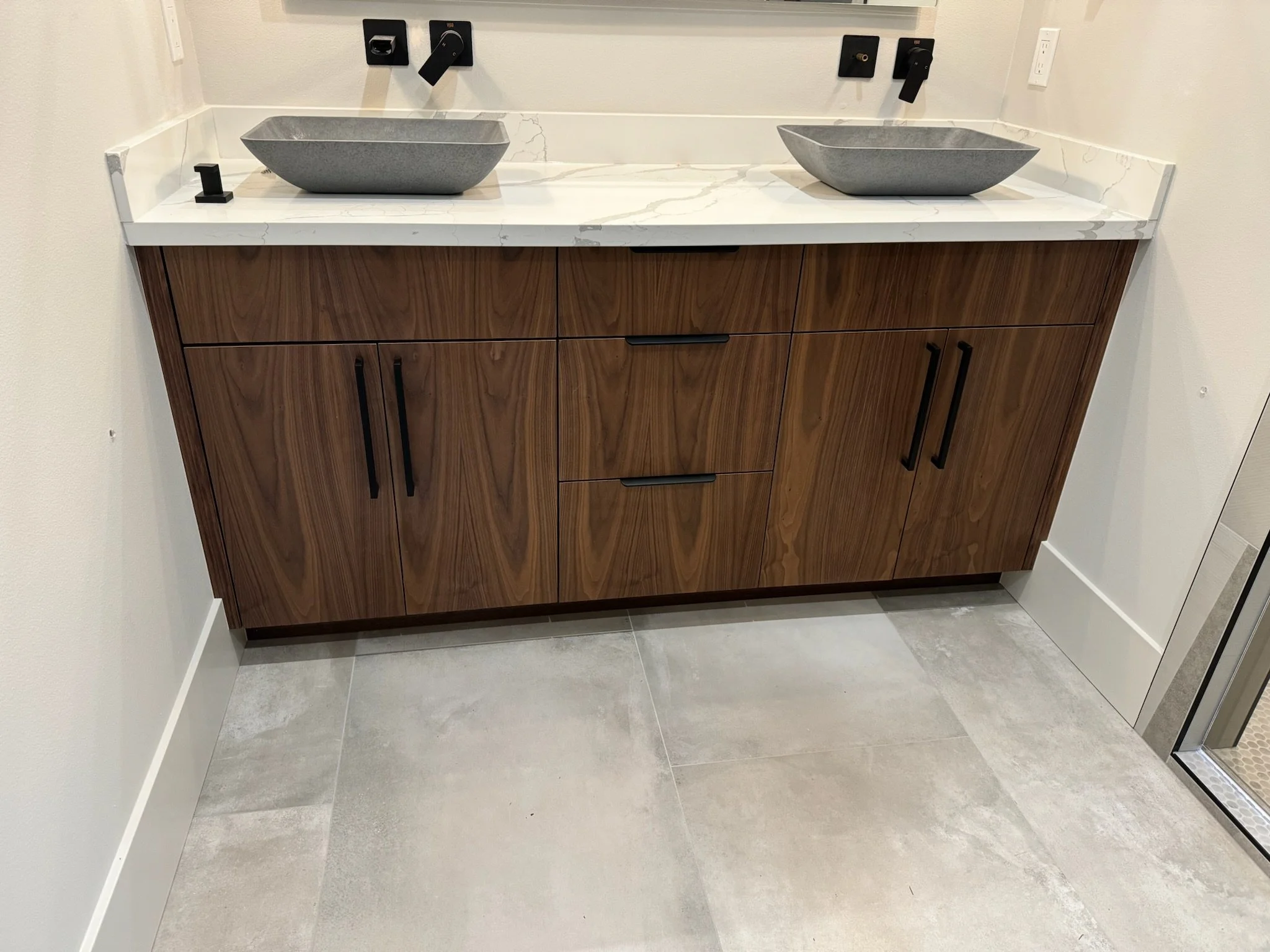 A bathroom vanity with a white marble countertop, two grey vessel sinks, and wooden cabinets with black handles.